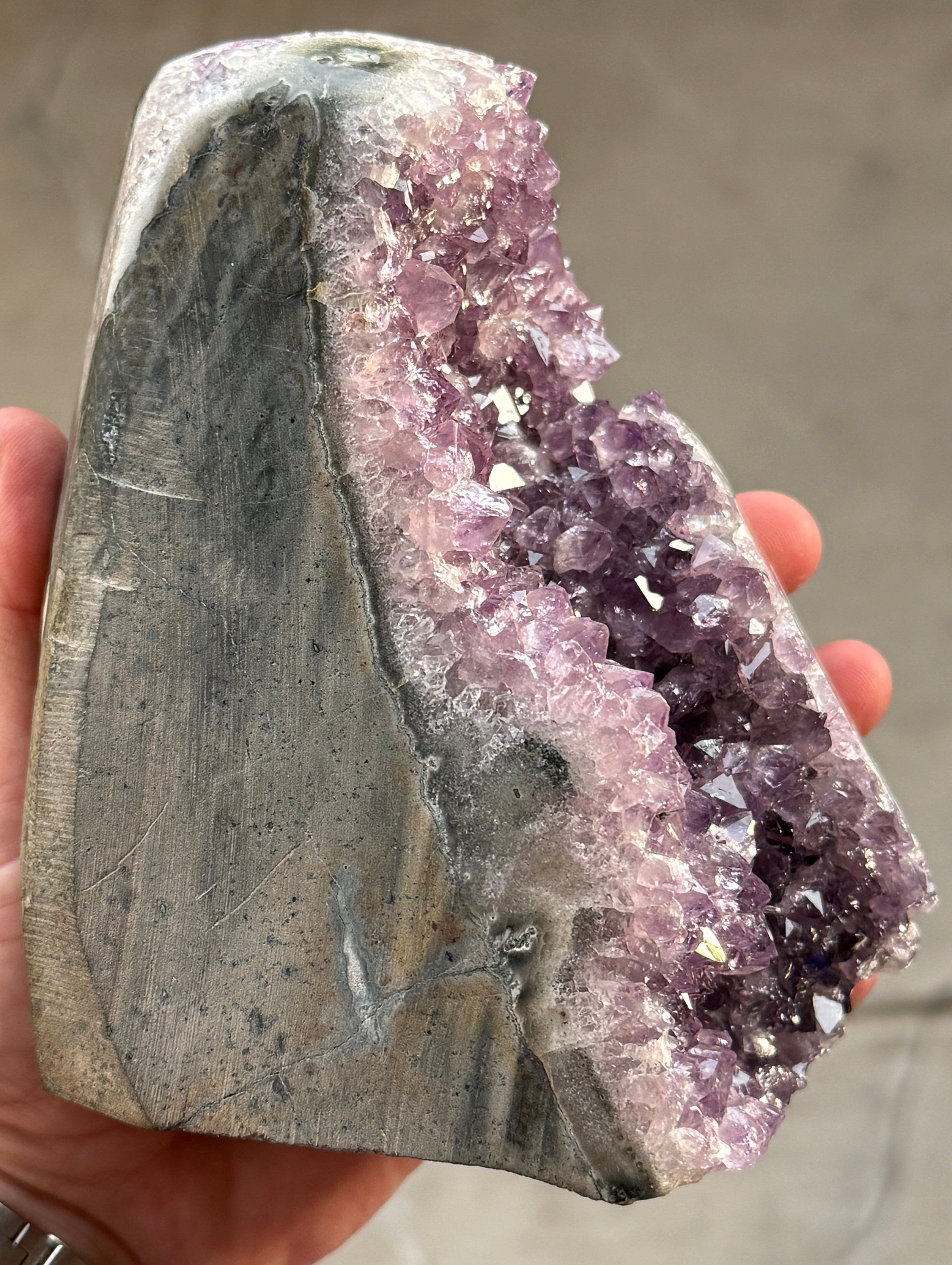 A large amethyst geode with a deep purple crystalline interior. The geode has a rough, natural exterior with a gray and white border. The crystals are densely packed and reflect light, creating a sparkling effect.