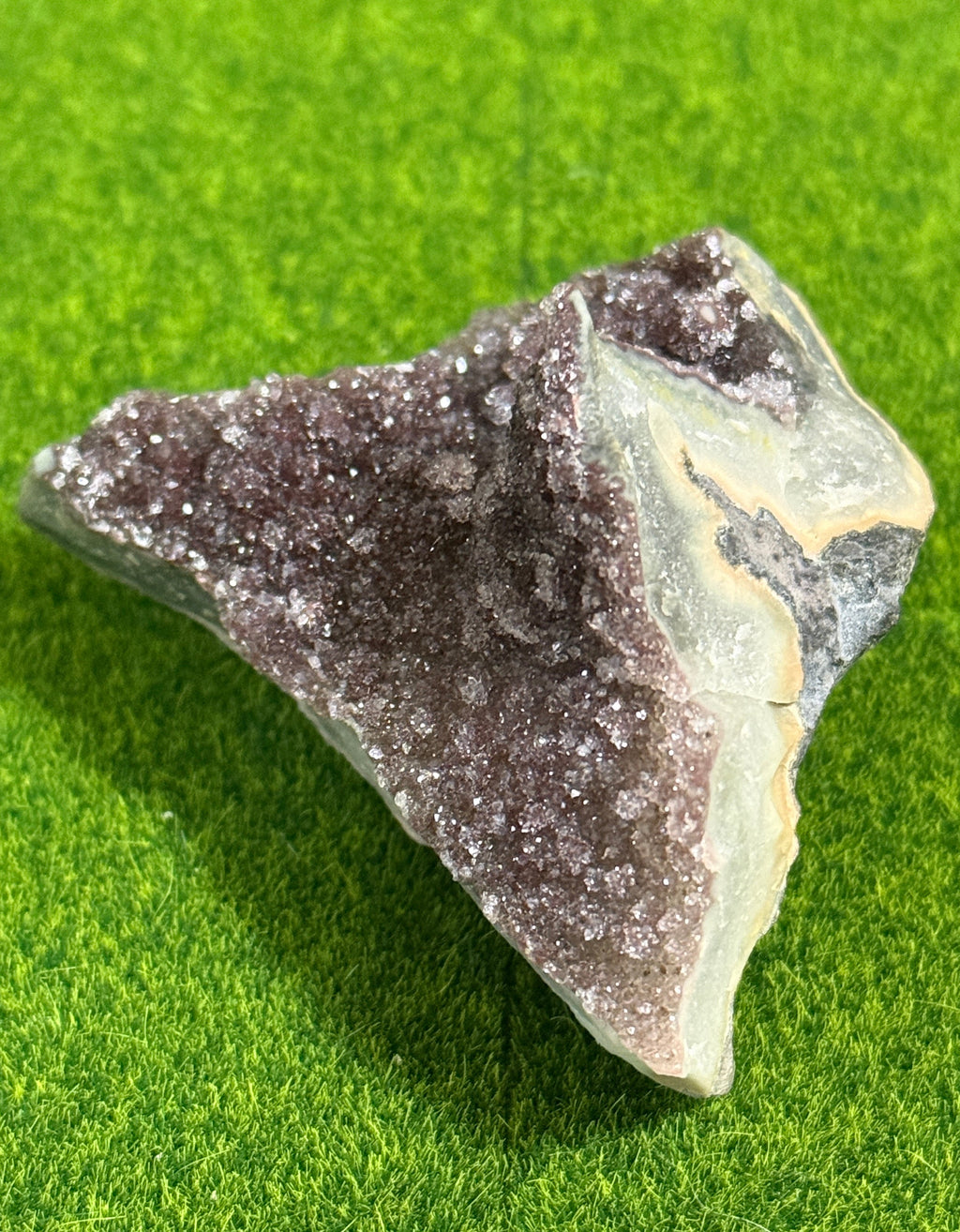 A natural amethyst geode specimen with a dark purple crystalline interior and a light green and beige exterior. The geode is displayed on a green surface.