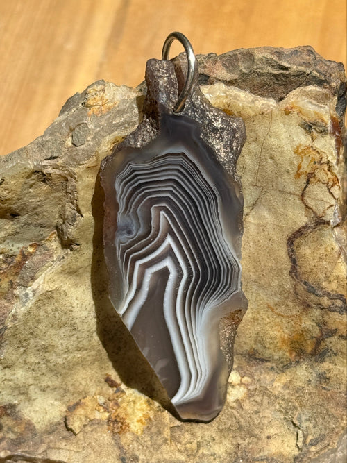 A polished agate stone pendant with concentric banding in gray, black, and white, attached to a silver split ring and displayed on a textured natural rock.