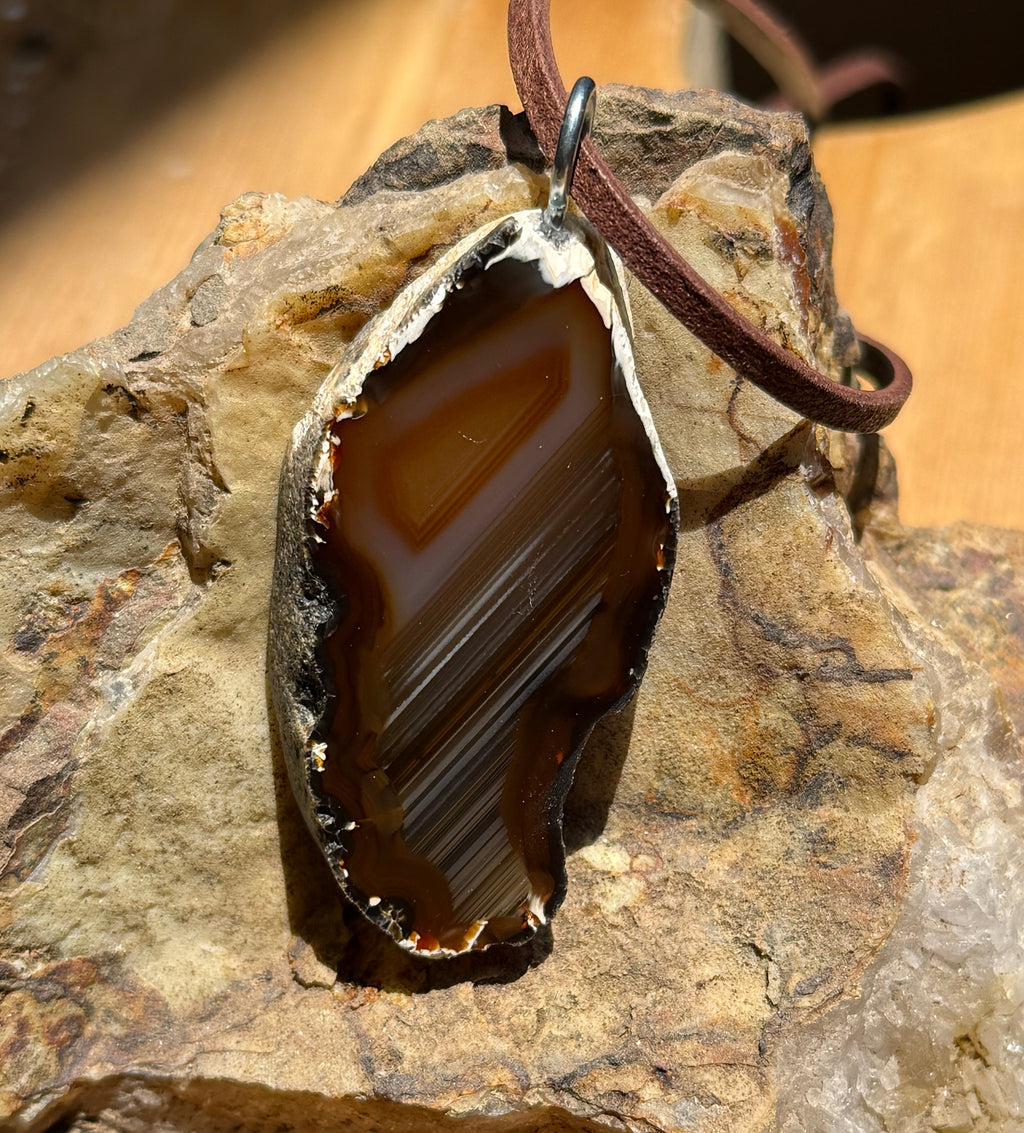 A raw agate stone pendant with a brown leather cord, resting on a textured natural rock against a wooden background.