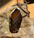 A raw agate stone pendant with a brown leather cord, resting on a textured natural rock against a wooden background.