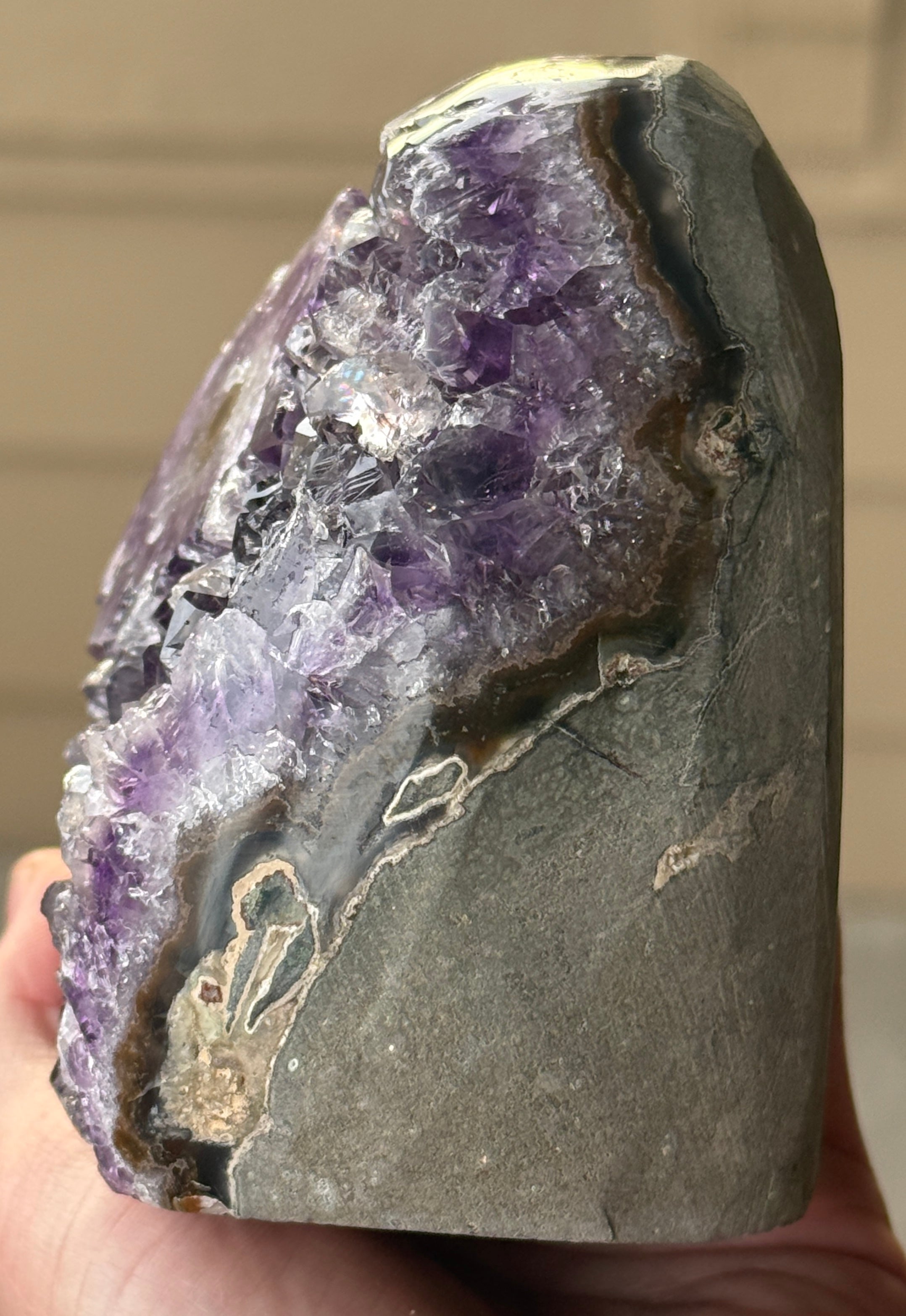 A polished amethyst geode with a deep purple hue. The crystal formation is visible, with numerous small, sparkling points. A dark, circular inclusion is present near the base, adding contrast to the overall color. The geode is a natural, decorative item.