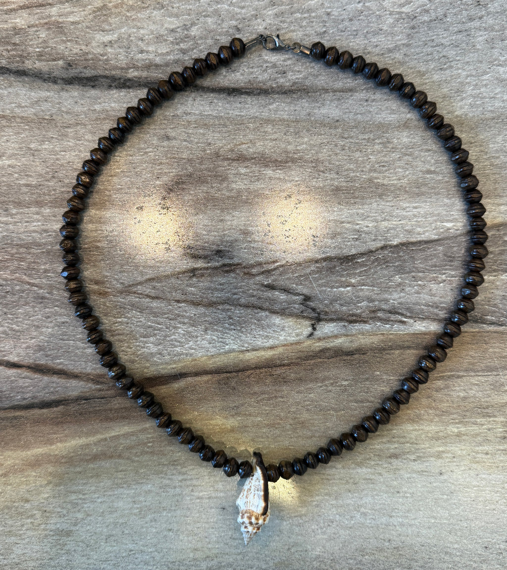A beaded necklace made of small dark brown beads with a conch shell pendant, displayed on a weathered wood background.