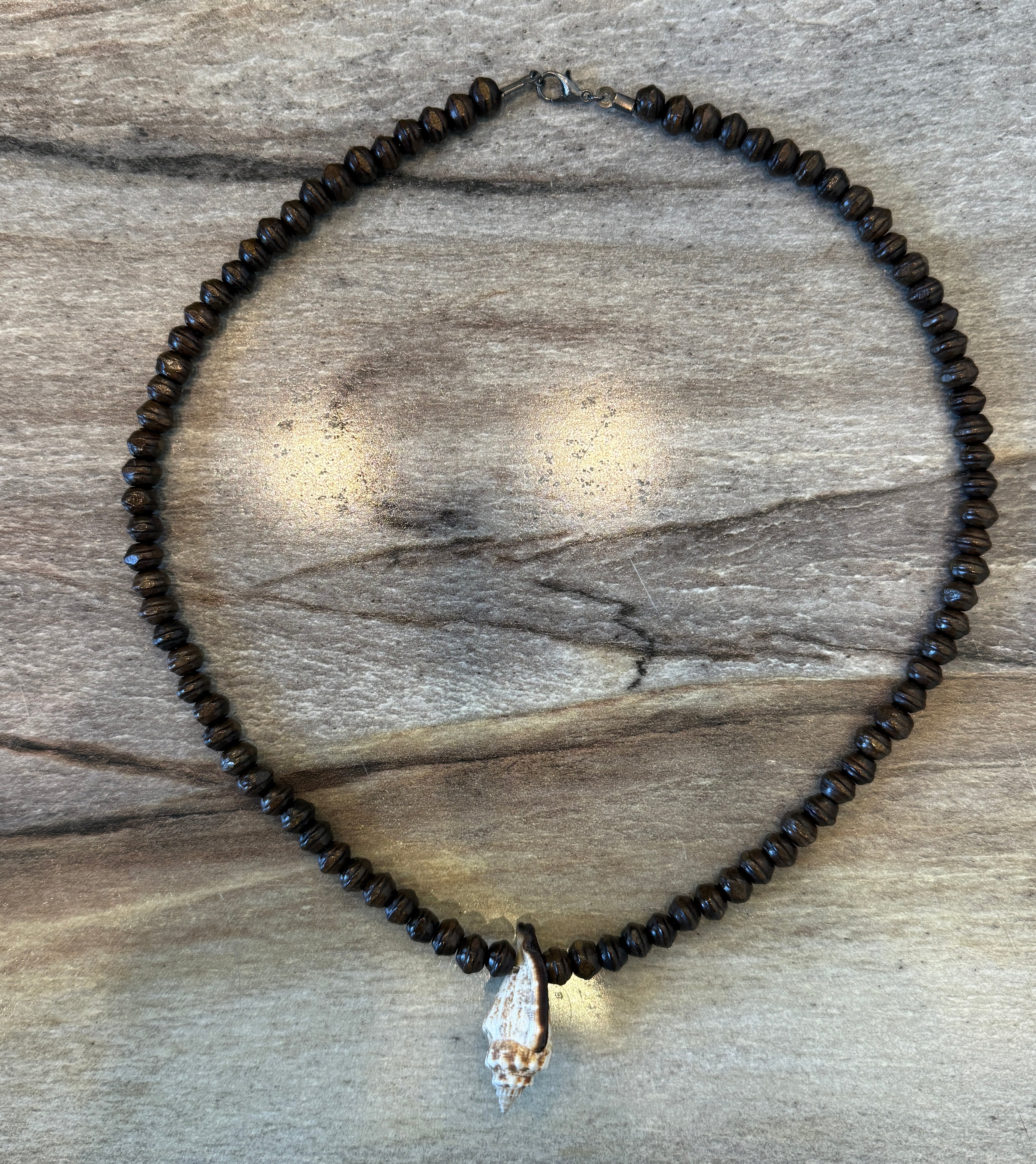 A beaded necklace made of small dark brown beads with a conch shell pendant, displayed on a weathered wood background.