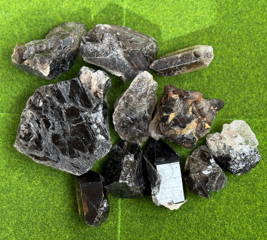 A collection of raw, uncut smoky quartz crystals. The crystals vary in size and shape, displaying shades of gray, brown, and black. They are arranged on a green surface, showcasing their natural, rough textures.