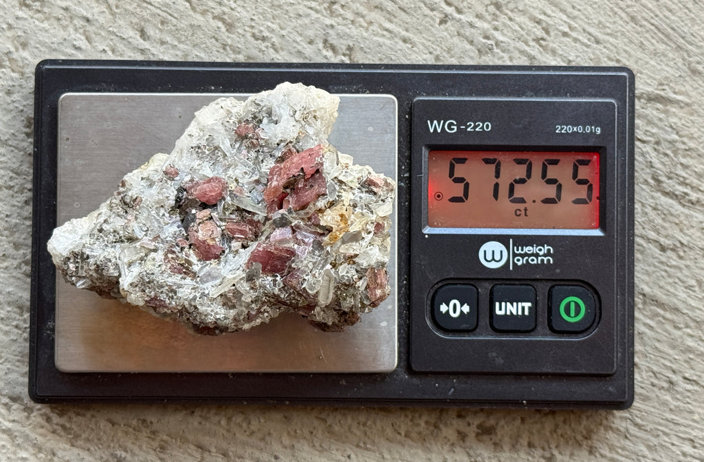 A raw mineral or rock specimen with pinkish-red inclusions sits on the weighing platform of a digital pocket scale displaying 572.55 grams.