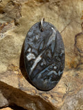 A polished oval black stone pendant with natural inclusions, displayed on a large textured rock over a wooden surface.