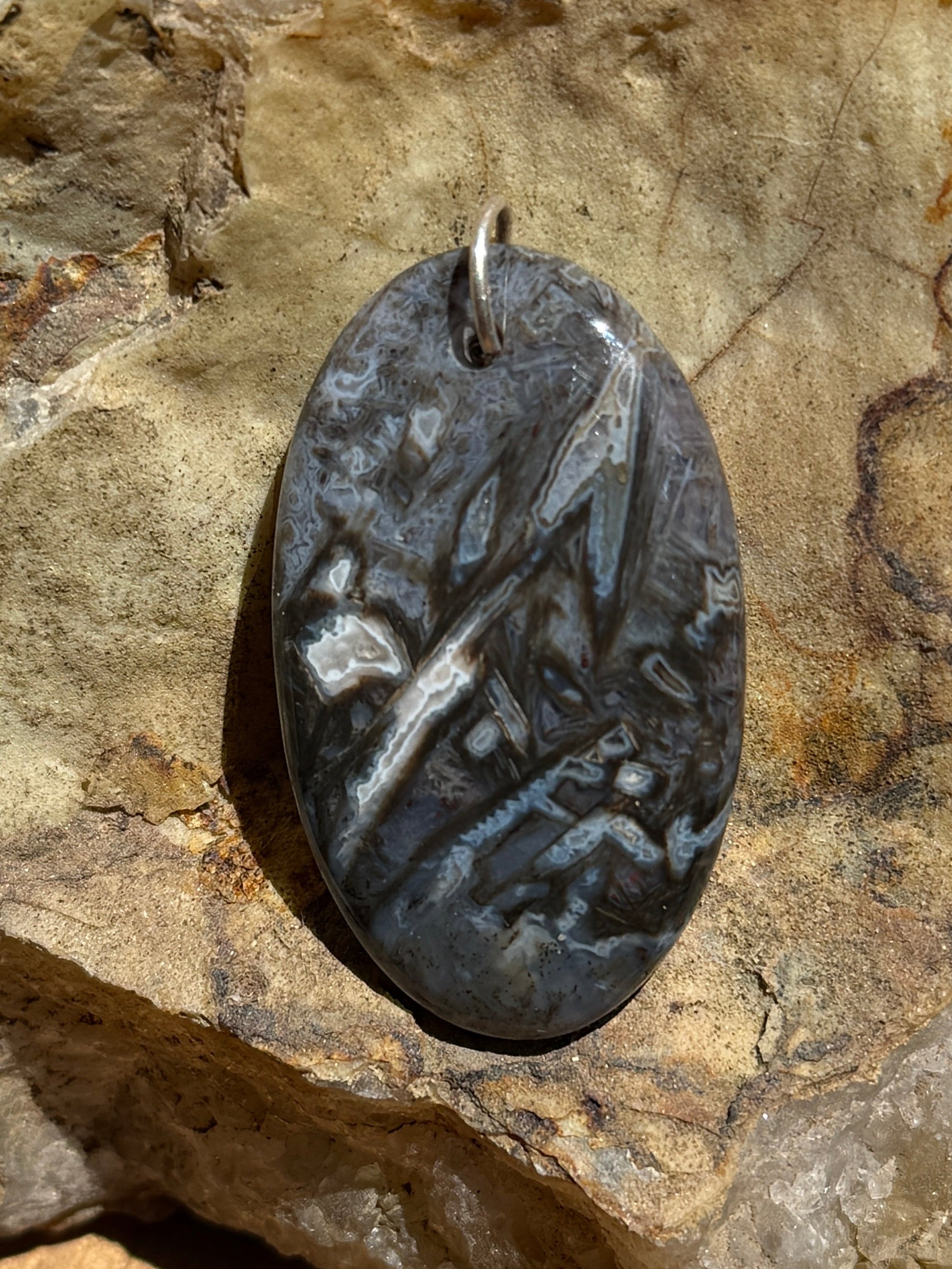 A polished oval black stone pendant with natural inclusions, displayed on a large textured rock over a wooden surface.