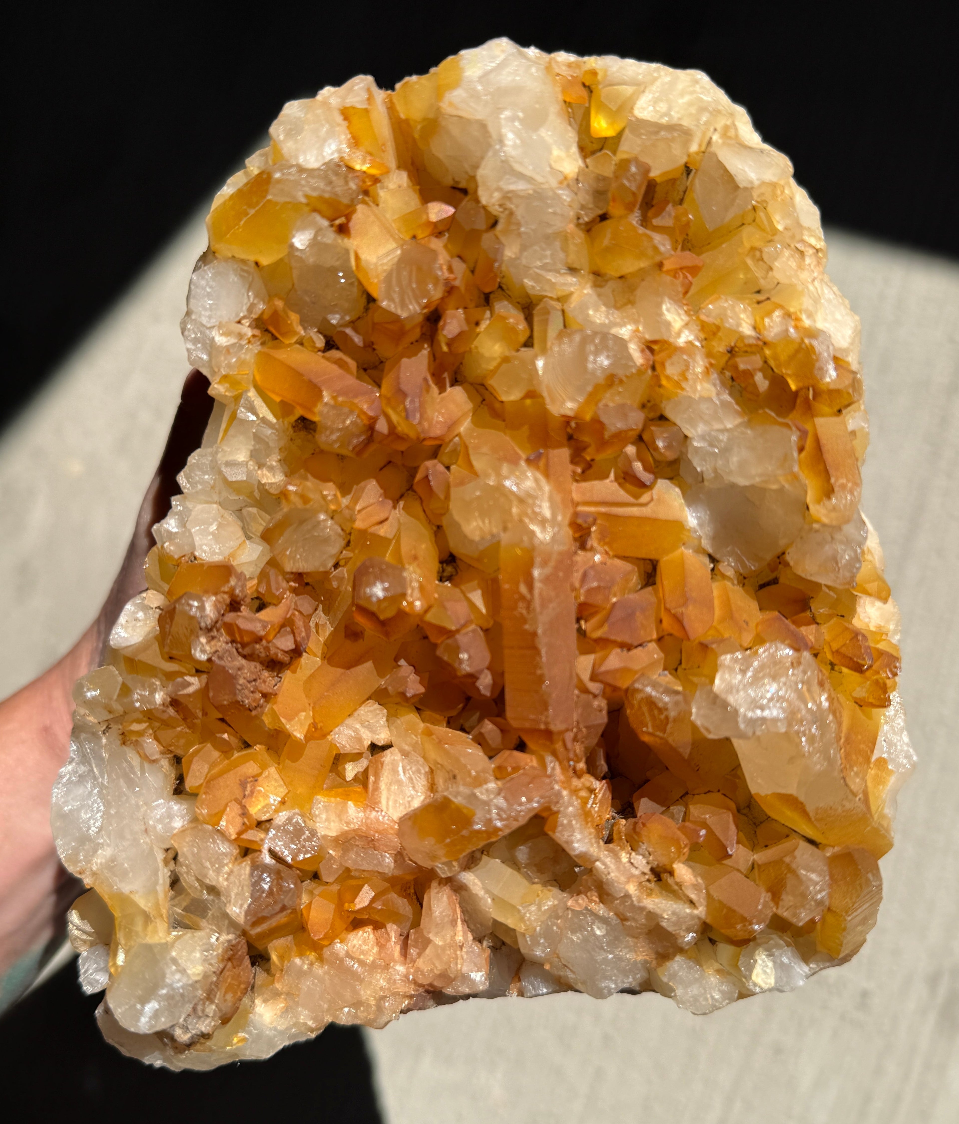 A hand holding a large crystal cluster specimen with prominent orange, amber, and translucent clear mineral formations against a dark and light background.