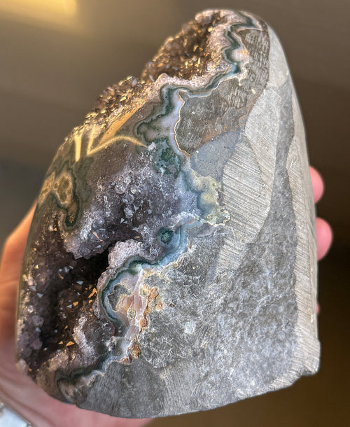A large, oval-shaped geode with a dark purple interior filled with sparkling crystals. The outer shell is a mottled gray-green with areas of teal and gold. The geode sits on a white surface. 
