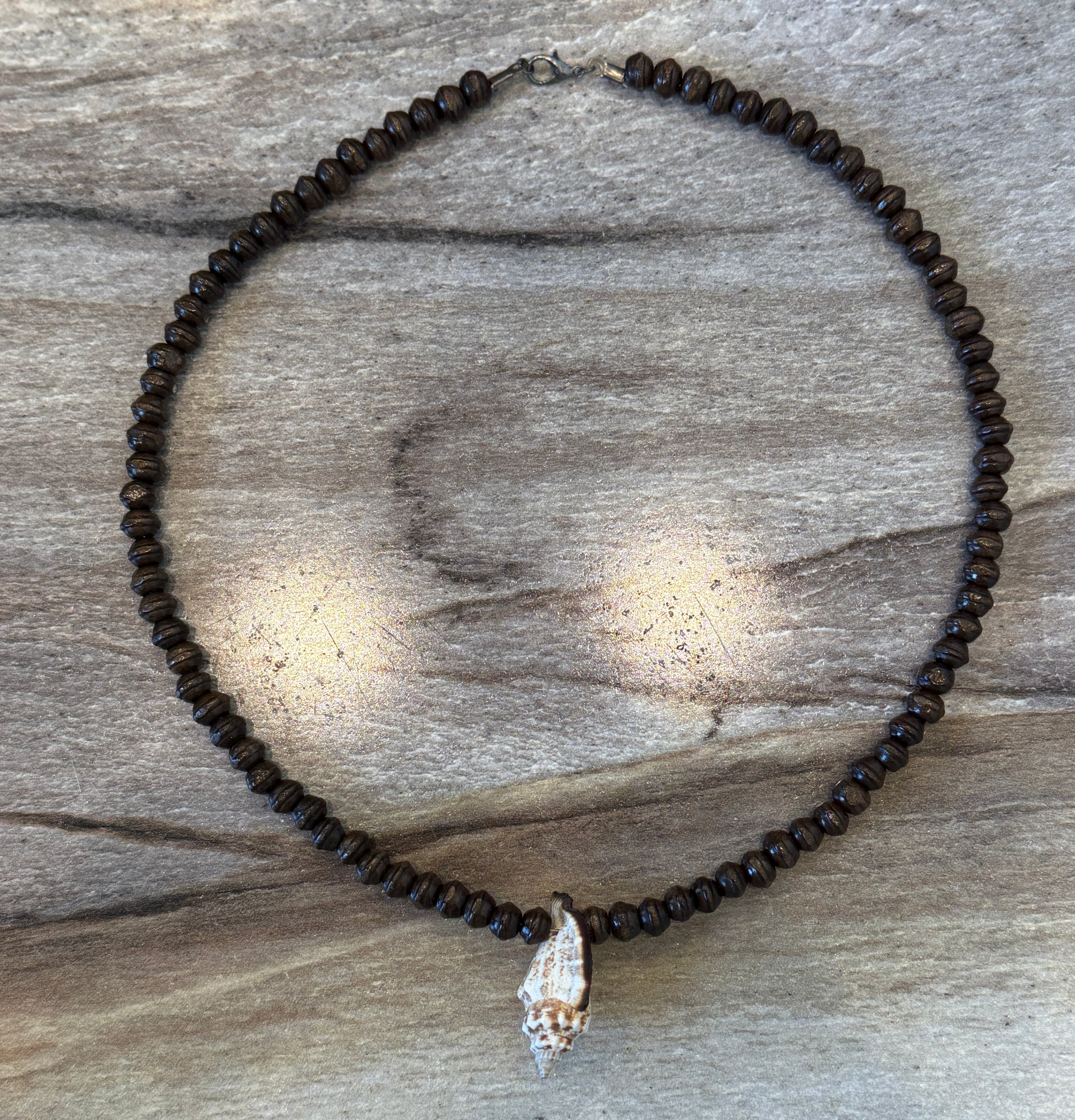 A beaded necklace made of small dark brown beads with a white seashell pendant, displayed on a weathered gray wood surface.