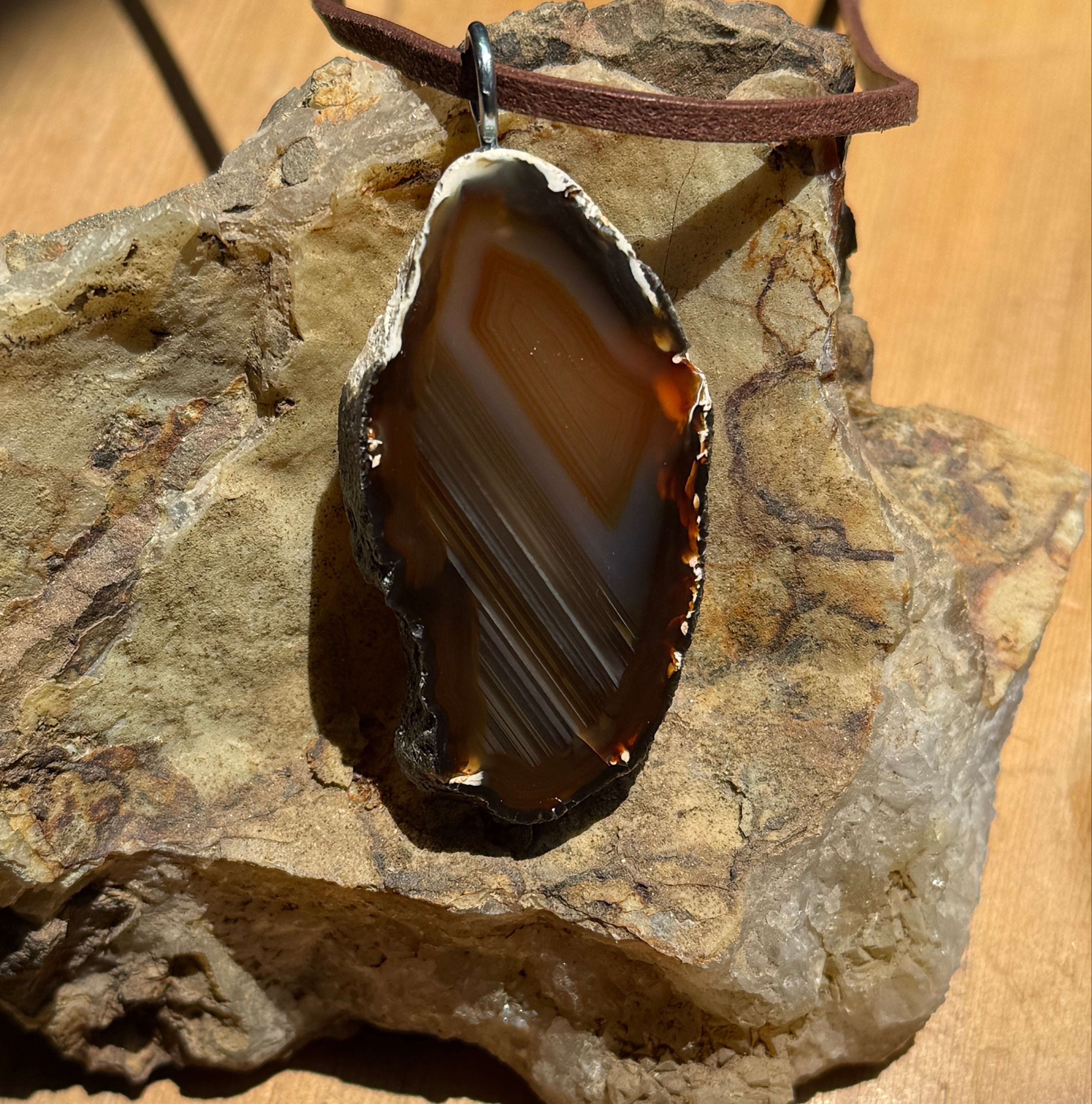 A polished agate slice pendant featuring concentric bands of amber, gold, and dark brown colors. The stone has a rough, natural outer edge and is fitted with a silver-toned metal bail