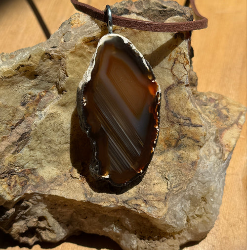 A polished agate slice pendant featuring concentric bands of amber, gold, and dark brown colors. The stone has a rough, natural outer edge and is fitted with a silver-toned metal bail