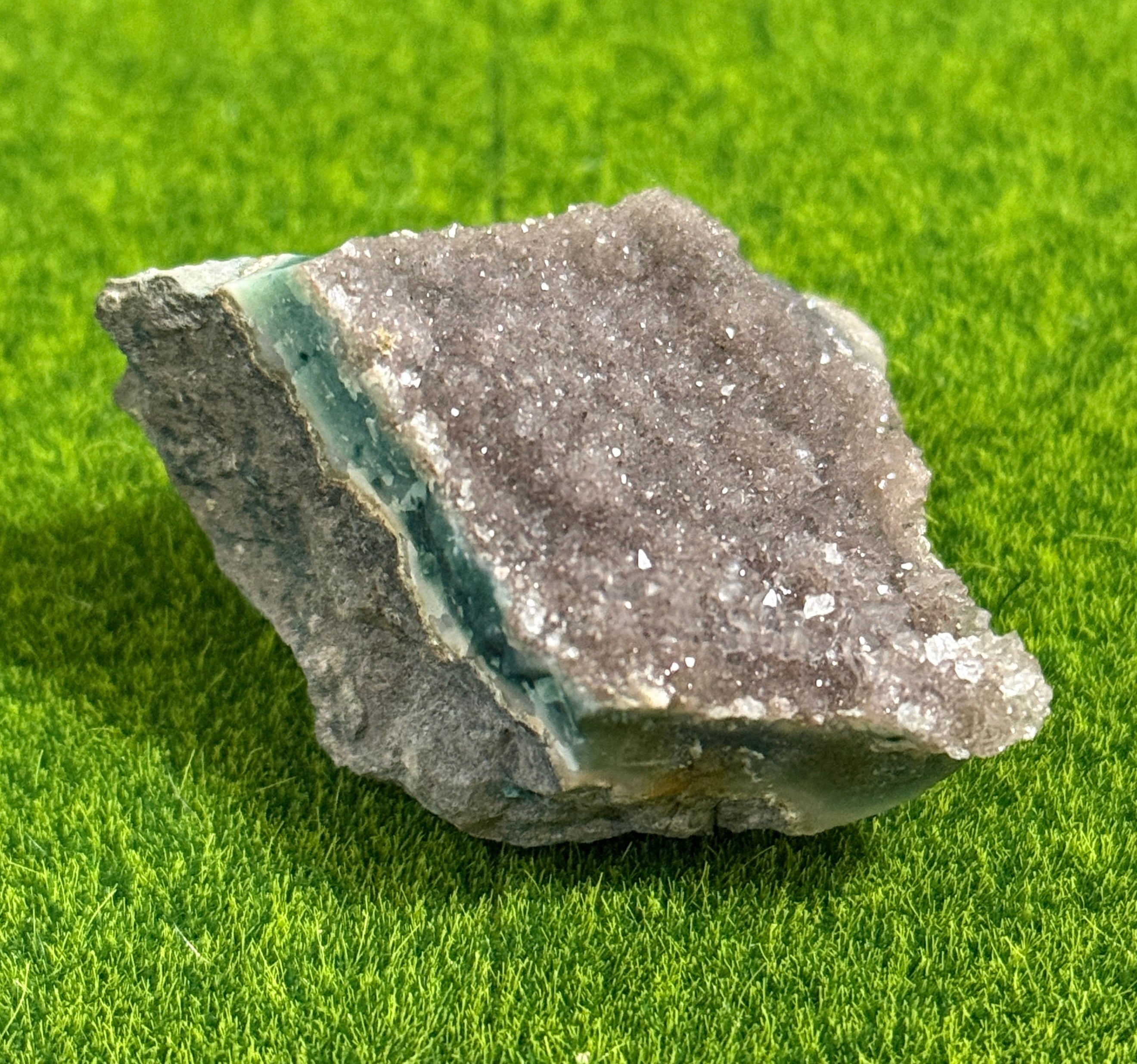 A rough-cut, natural amethyst geode with a crystalline surface. The geode features a band of teal-colored mineral deposits. The stone is set against a bright green background, highlighting its natural colors and textures.
