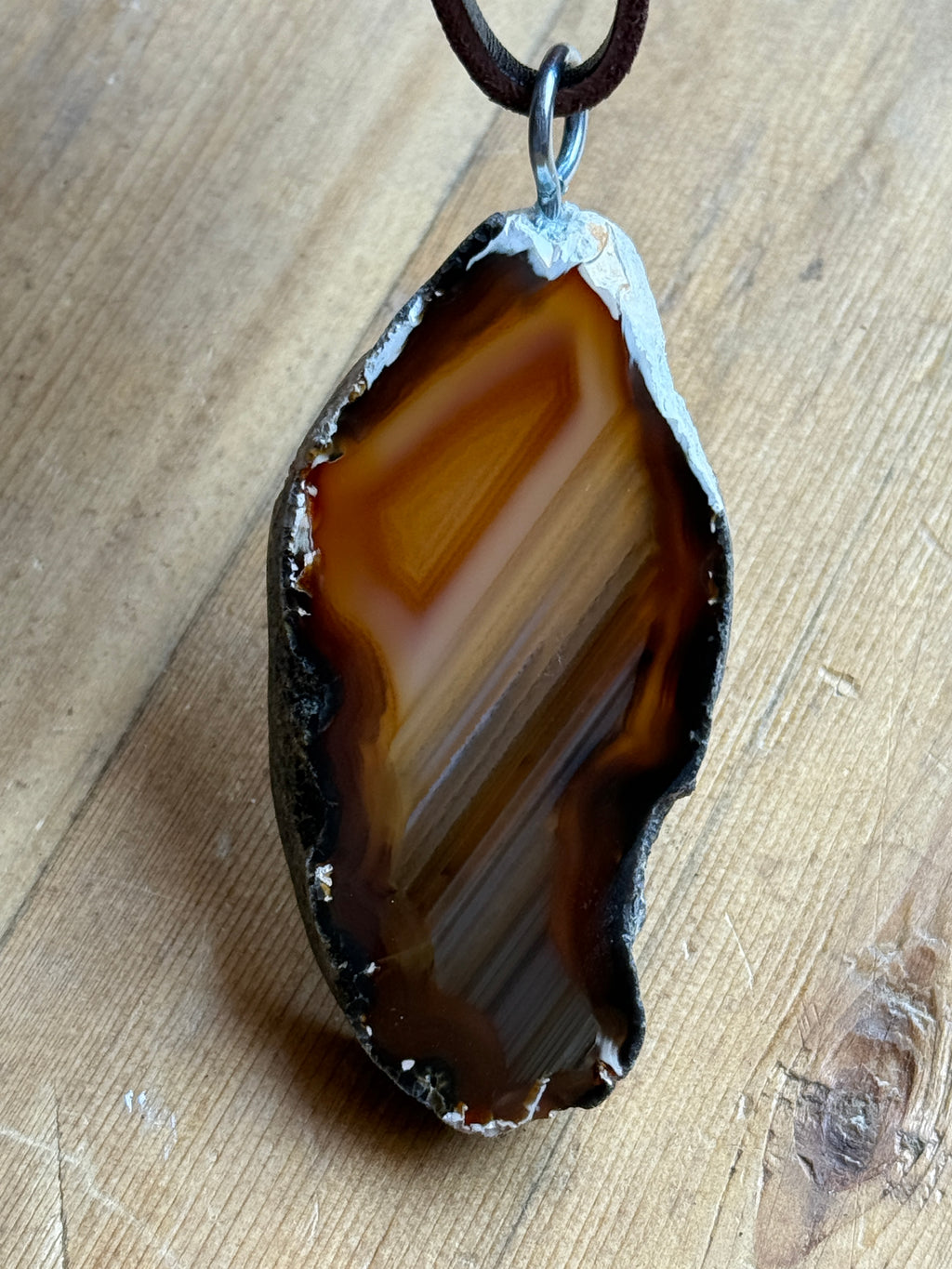 A natural agate stone pendant with a rough, polished edge and a dark brown leather loop attached, displayed on a light wood surface.