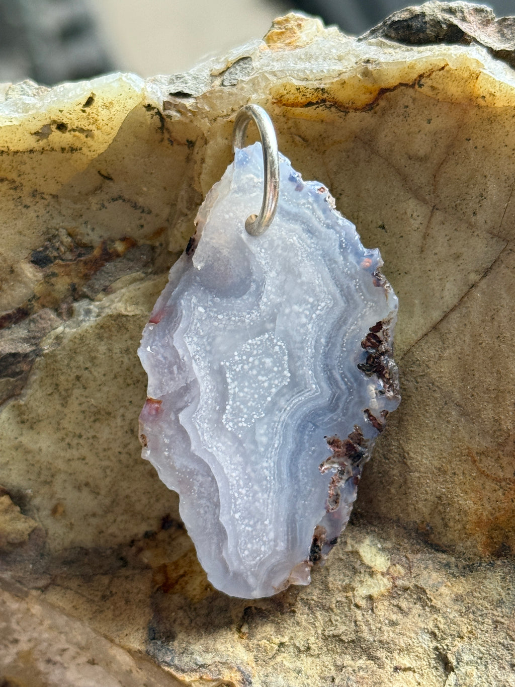 Blue agate geode slice pendant with a silver jump ring, displayed on a textured natural rock background.