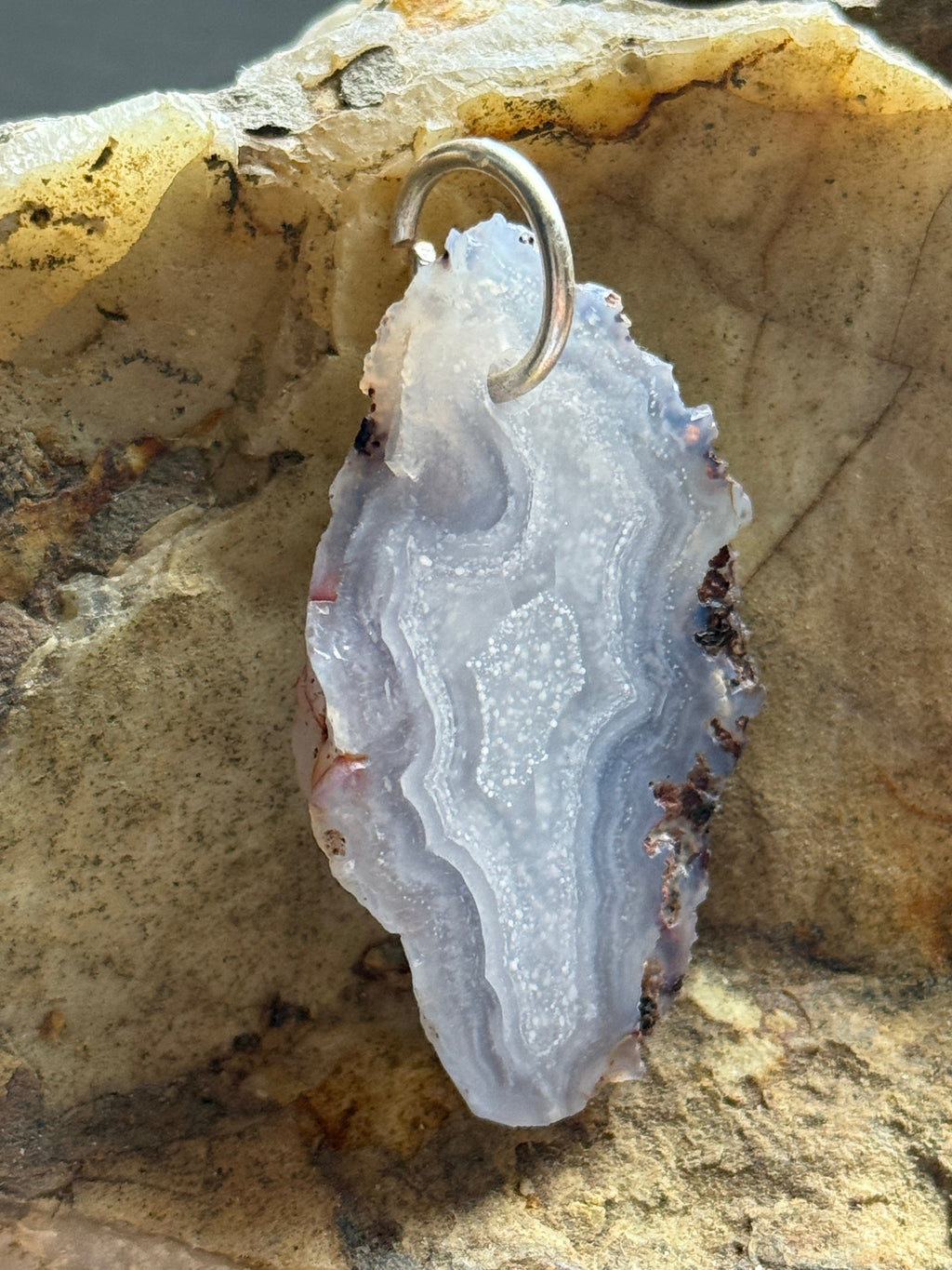A polished agate geode slice pendant with a silver bail, displayed on a textured natural rock surface.