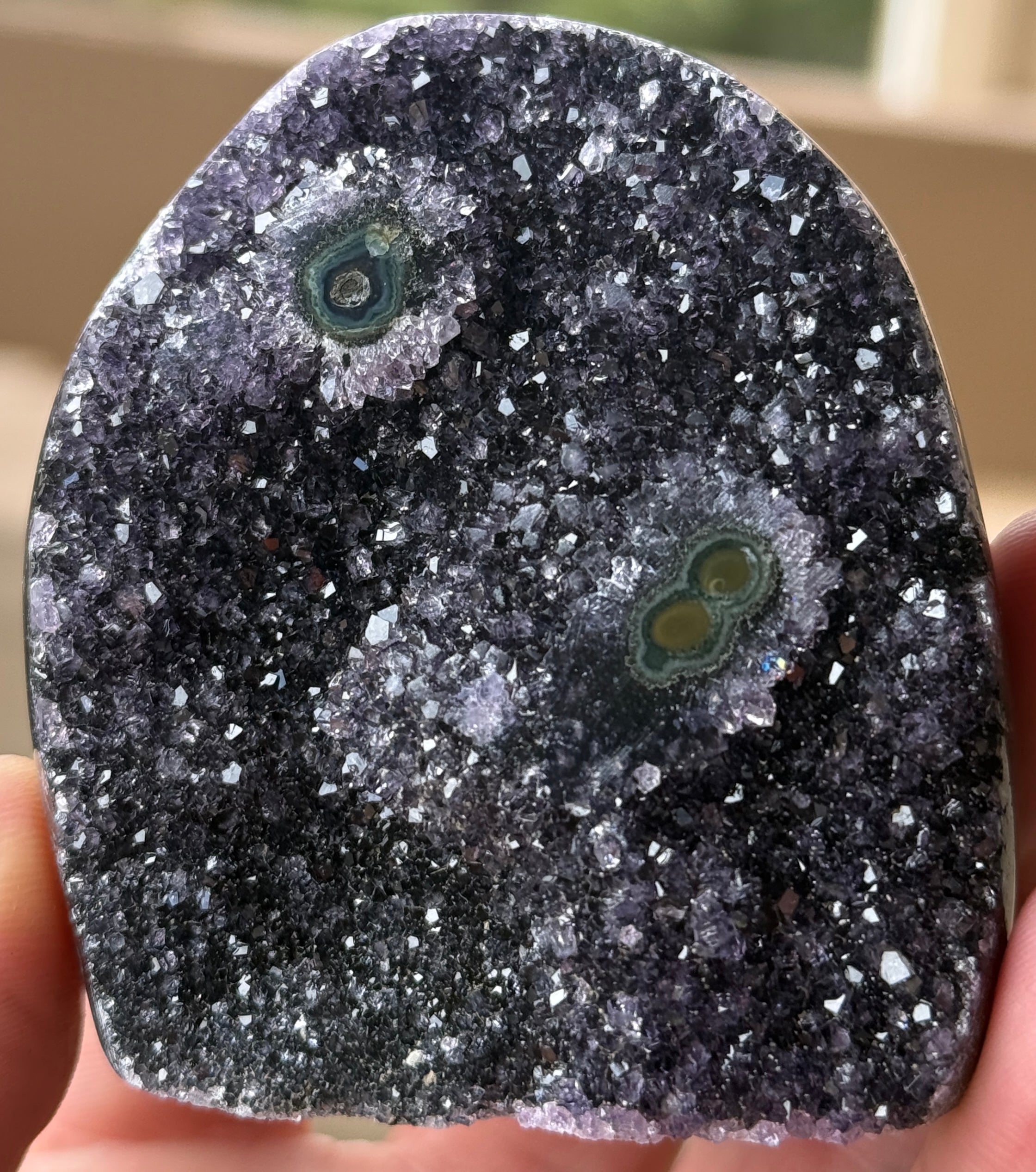A polished amethyst geode with a dark purple crystalline interior. The geode features two distinct eye-like formations with concentric rings of green and brown. The exterior is a smooth, rounded shape.