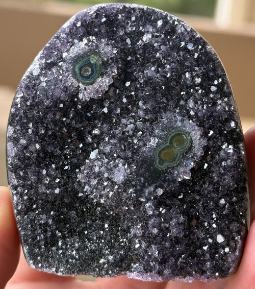 A polished amethyst geode with a dark purple crystalline interior. The geode features two distinct eye-like formations with concentric rings of green and brown. The exterior is a smooth, rounded shape.