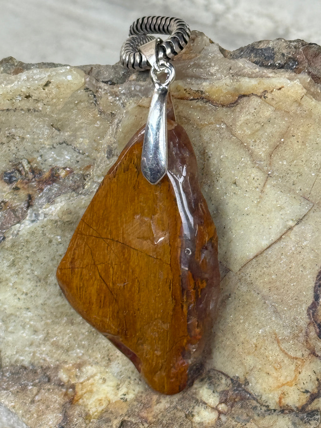 A polished brown stone pendant with a silver-toned bail and chain, displayed on a textured beige and gray stone surface.