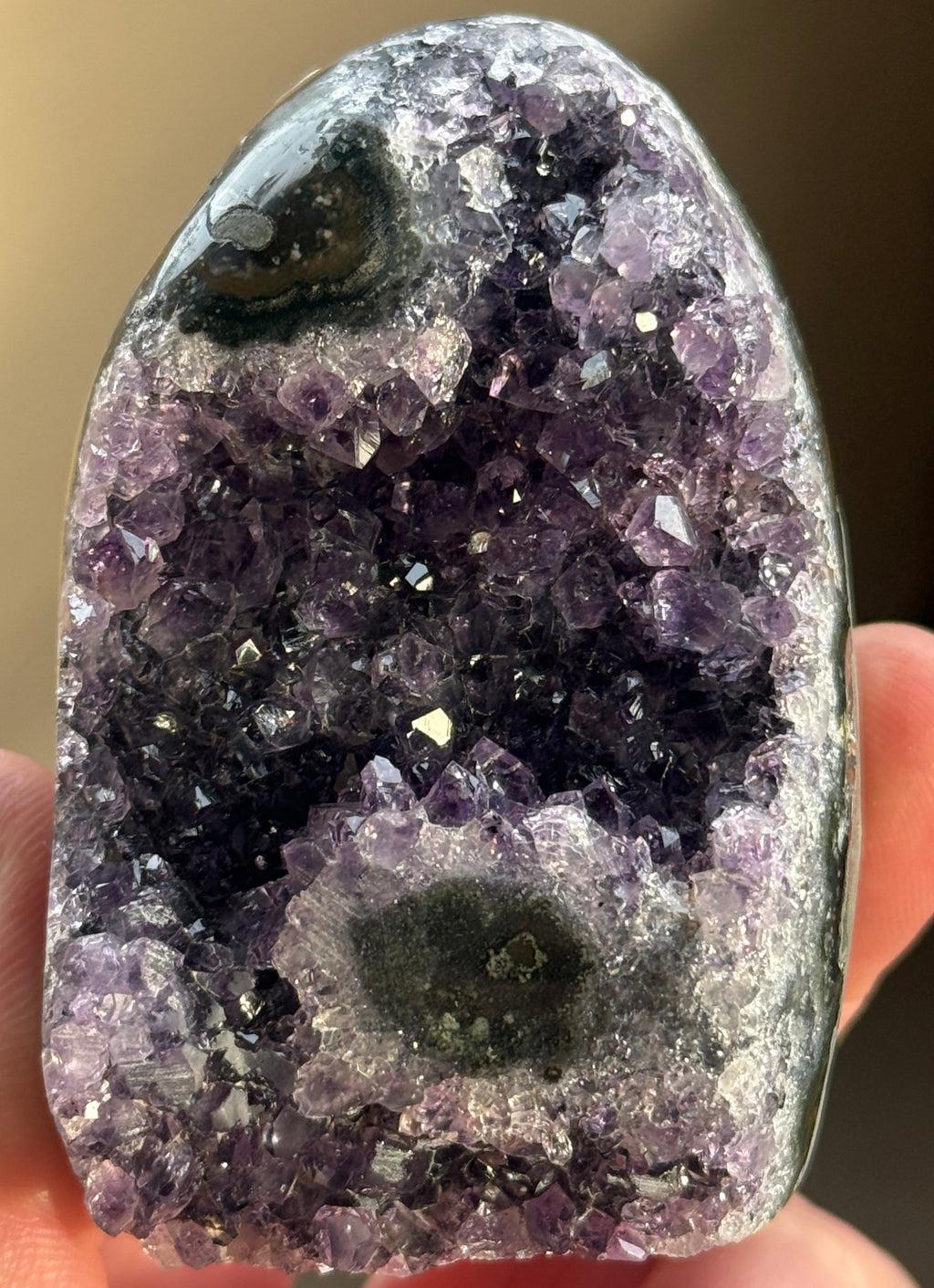A polished amethyst geode with a deep purple hue. The crystal formation is visible, with numerous small, sparkling points. A dark, circular inclusion is present near the base, adding contrast to the overall color. The geode is a natural, decorative item.