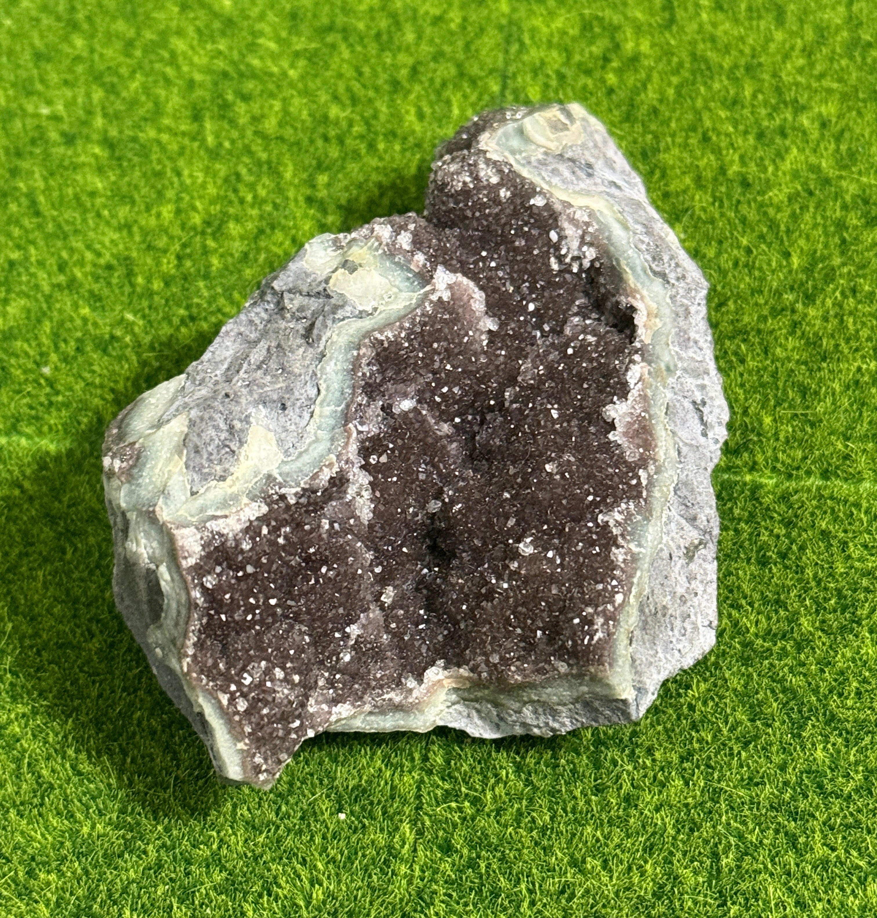 A natural geode with a dark purple crystal center and a gray and green outer layer. The geode has a rough, textured surface and is displayed on a green background.