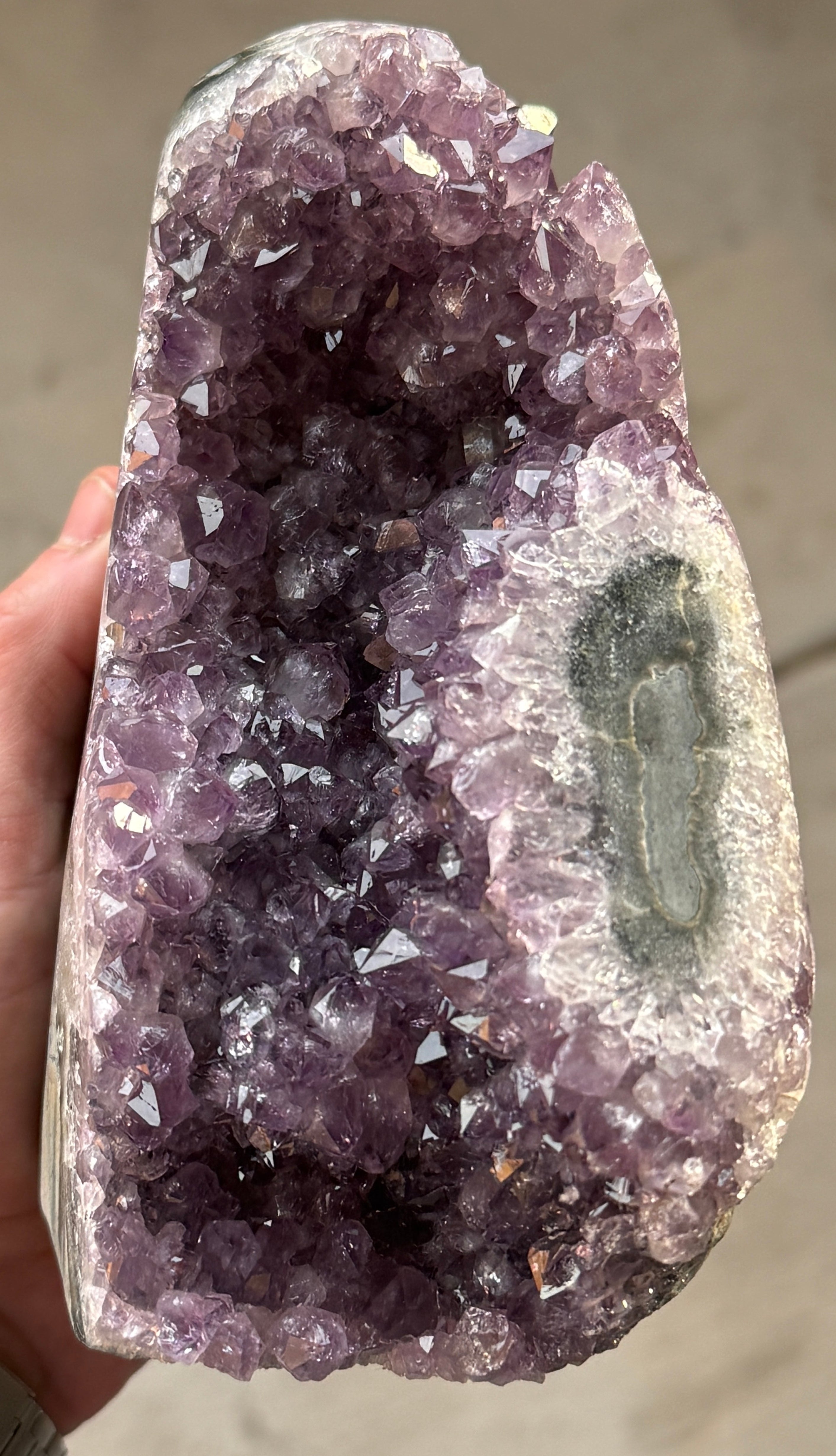 A large amethyst geode with a deep purple crystalline interior. The geode has a rough, natural exterior with a gray and white border. The crystals are densely packed and reflect light, creating a sparkling effect.