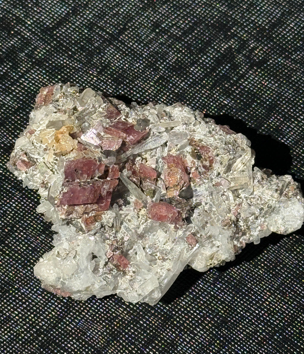 A raw mineral specimen with clear quartz crystals and reddish-brown metallic inclusions, resting on a black textured mesh background.