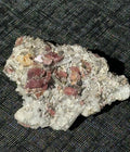 A raw mineral specimen with clear quartz crystals and reddish-brown metallic inclusions, resting on a black textured mesh background.