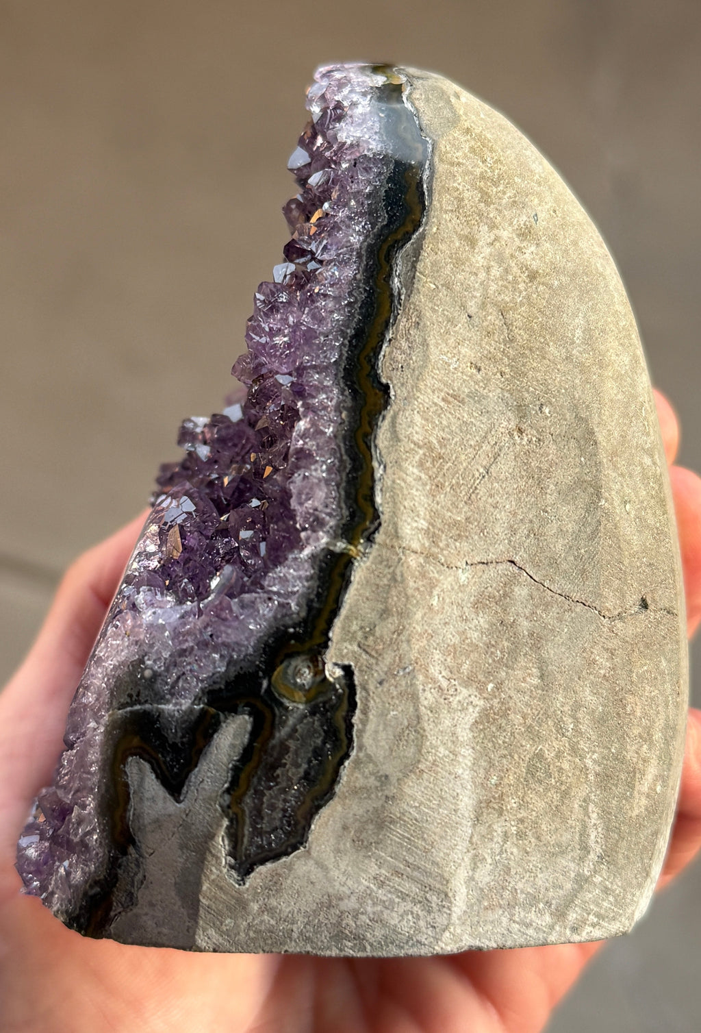 Side view of a natural amethyst geode with a deep purple crystal interior. The geode has a rounded shape and a rough, textured exterior. A small, circular formation with green and yellow hues is visible within the crystal cluster.
