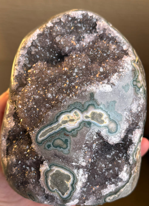 A large, oval-shaped geode with a dark purple interior filled with sparkling crystals. The outer shell is a mottled gray-green with areas of teal and gold. The geode sits on a white surface.