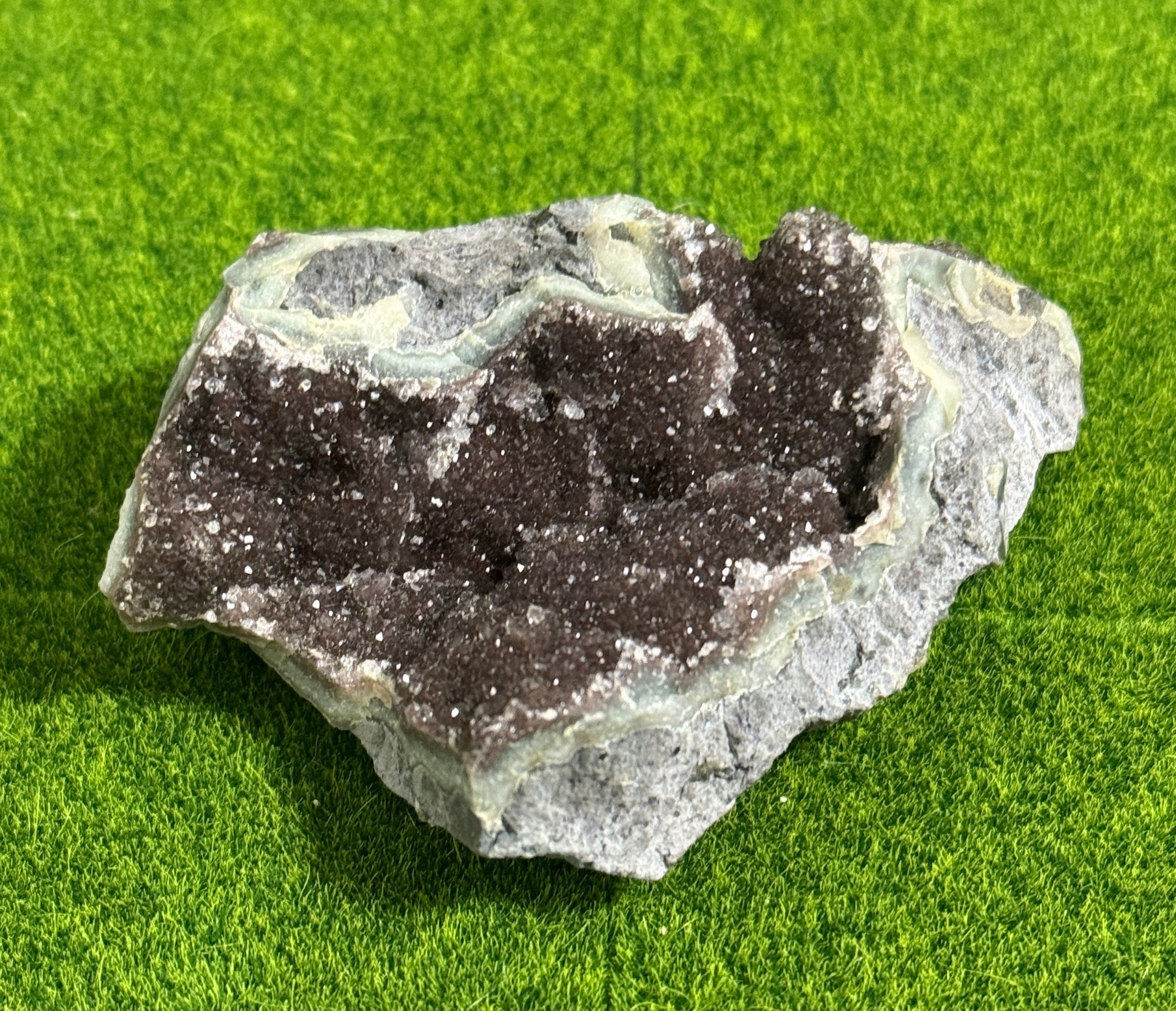 A rough-hewn, natural geode specimen. The exterior is a mottled gray and pale green, framing a cavity filled with sparkling, dark crystals. The geode rests on a bright green surface.