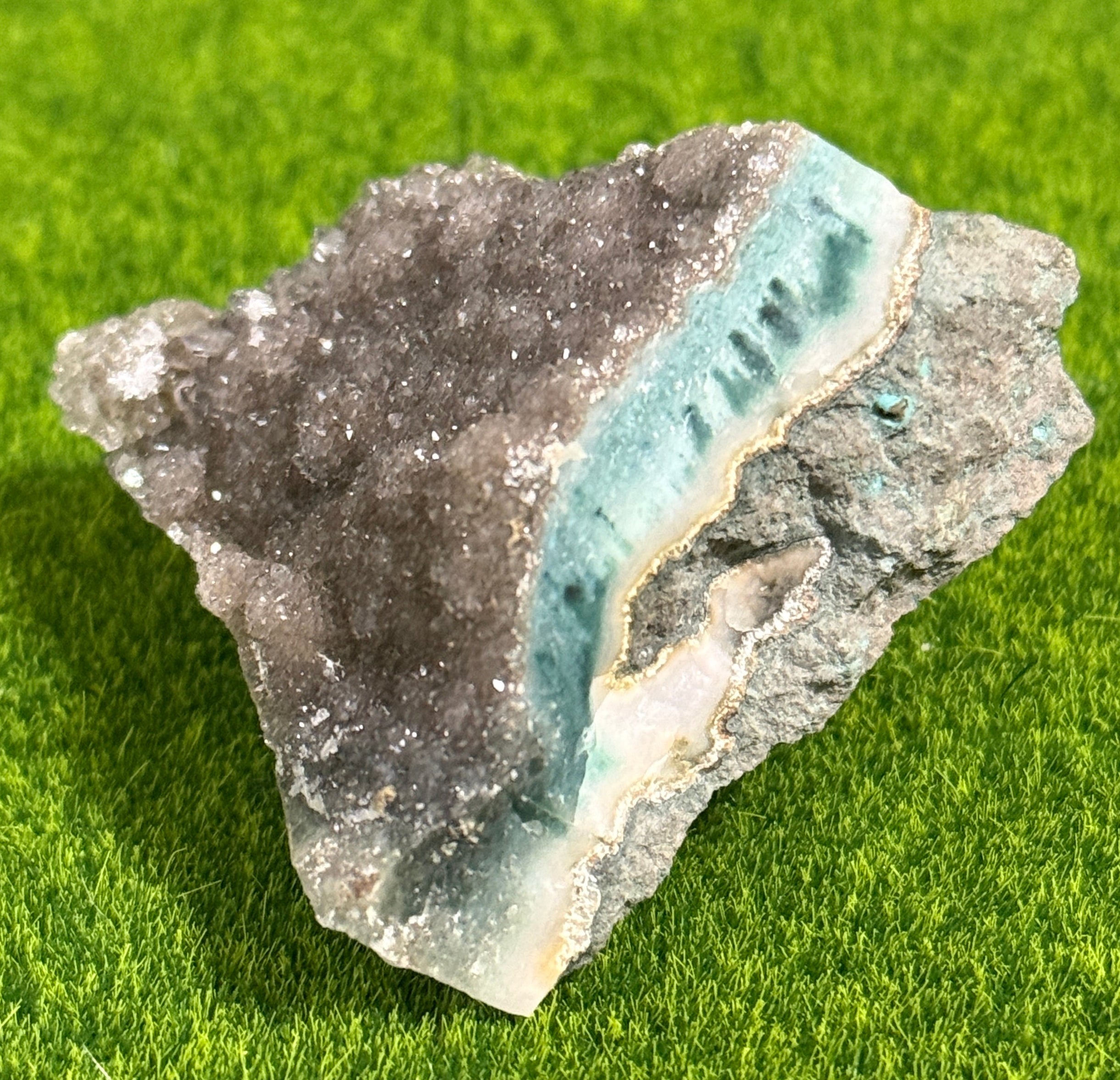 A natural mineral specimen featuring a mix of colors. The stone has a dark gray, crystalline exterior with a band of light blue and white in the center. The mineral sits on a green surface.