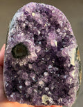 A polished amethyst geode with a deep purple hue. The crystal formation is visible, with numerous small, sparkling points. A dark, circular inclusion is present near the base, adding contrast to the overall color. The geode is a natural, decorative item.