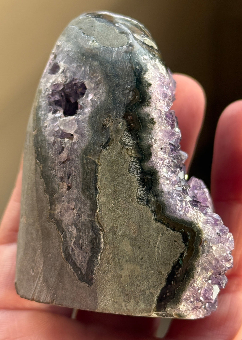 A polished amethyst geode with a deep purple hue. The crystal formation is visible, with numerous small, sparkling points. A dark, circular inclusion is present near the base, adding contrast to the overall color. The geode is a natural, decorative item.