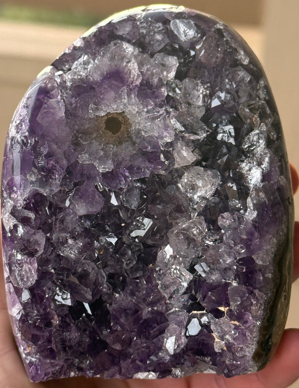 A polished amethyst geode with a deep purple hue. The crystal formation is visible, with numerous small, sparkling points. A dark, circular inclusion is present near the base, adding contrast to the overall color. The geode is a natural, decorative item.