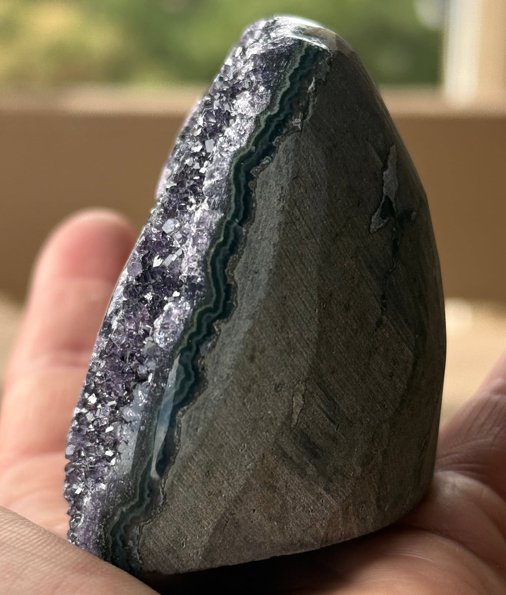 A polished amethyst geode with a dark purple crystalline interior. The geode features two distinct eye-like formations with concentric rings of green and brown. The exterior is a smooth, rounded shape.