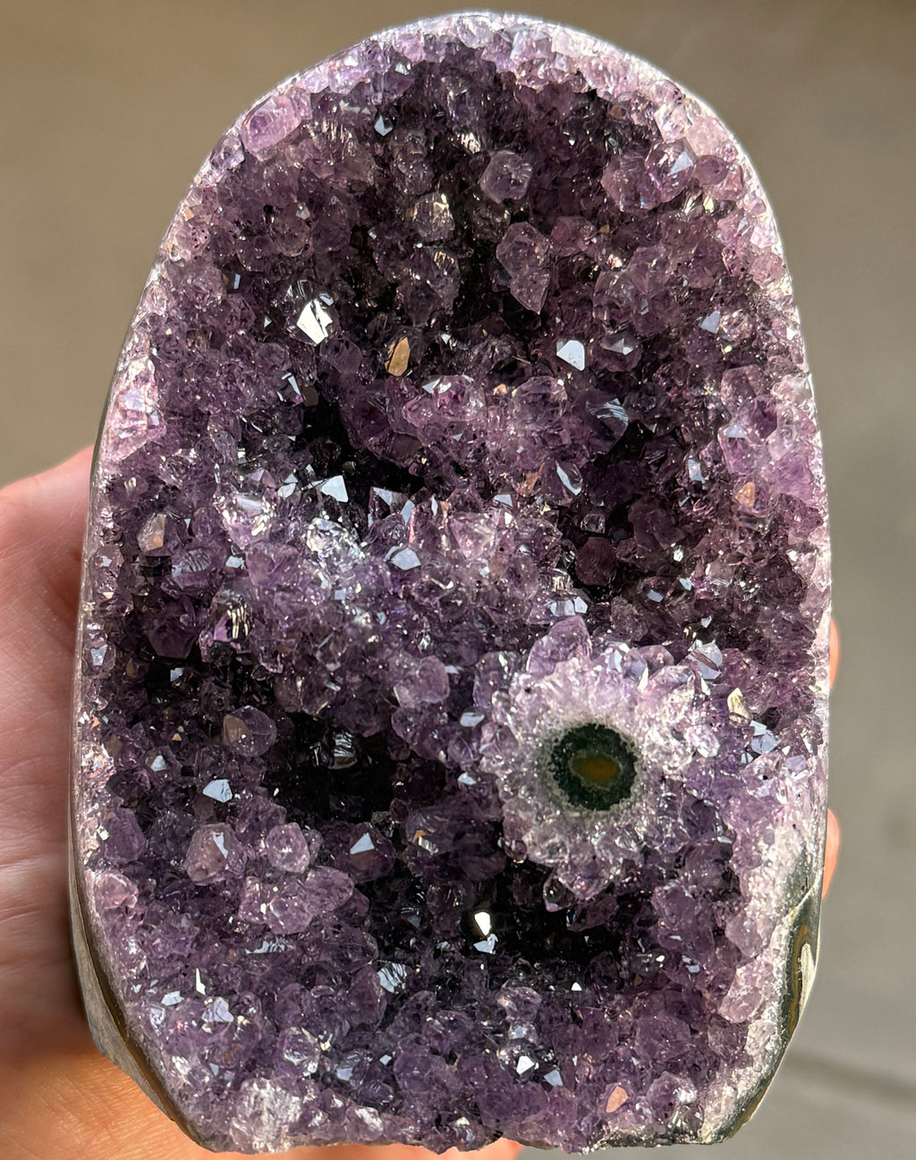 A natural amethyst geode with a deep purple crystal interior. The geode has a rounded shape and a rough, textured exterior. A small, circular formation with green and yellow hues is visible within the crystal cluster.