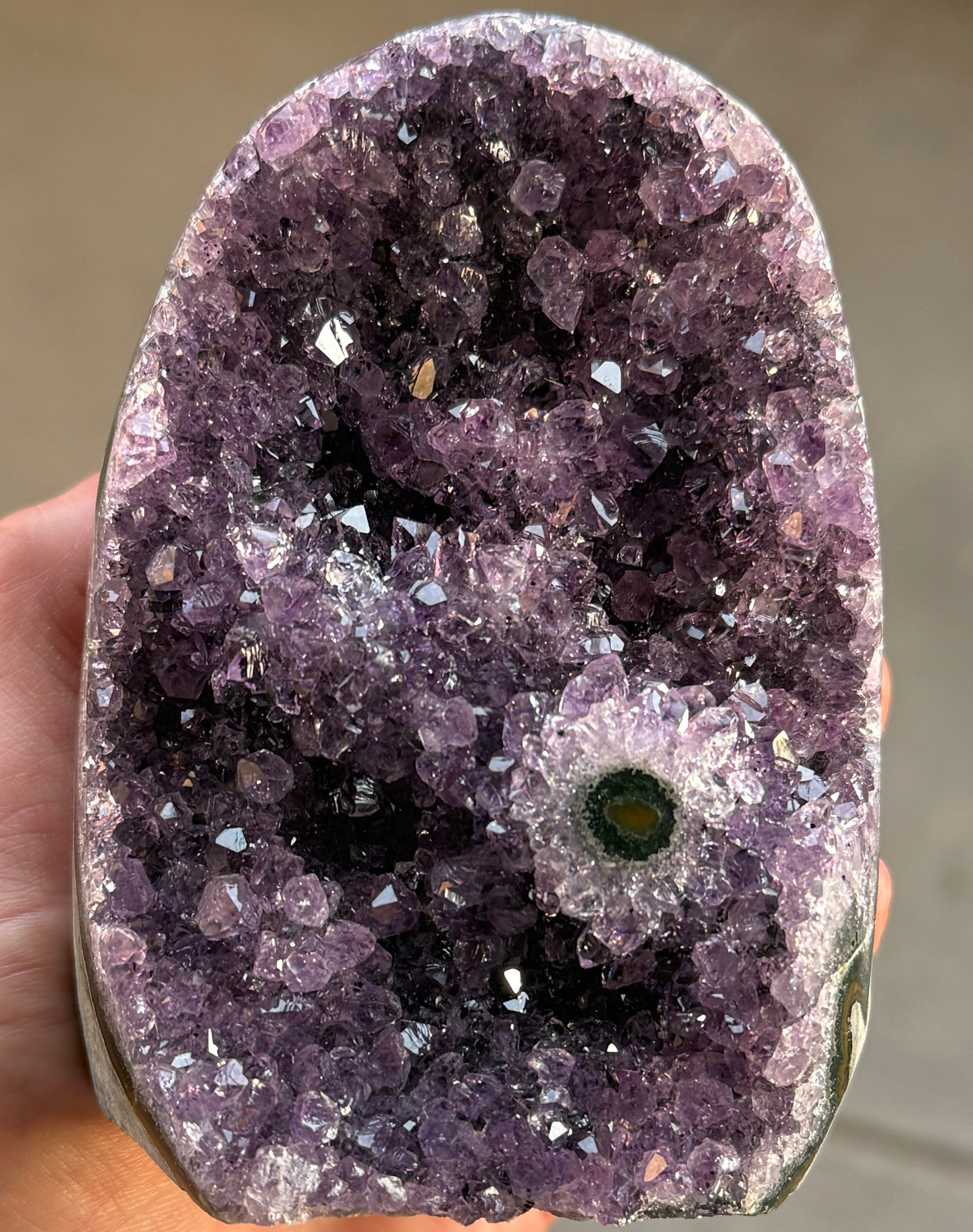 A natural amethyst geode with a deep purple crystal interior. The geode has a rounded shape and a rough, textured exterior. A small, circular formation with green and yellow hues is visible within the crystal cluster.