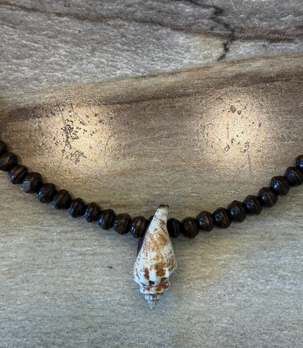 A beaded necklace with dark brown wooden beads and a natural conch shell pendant, displayed on a textured gray wood surface.