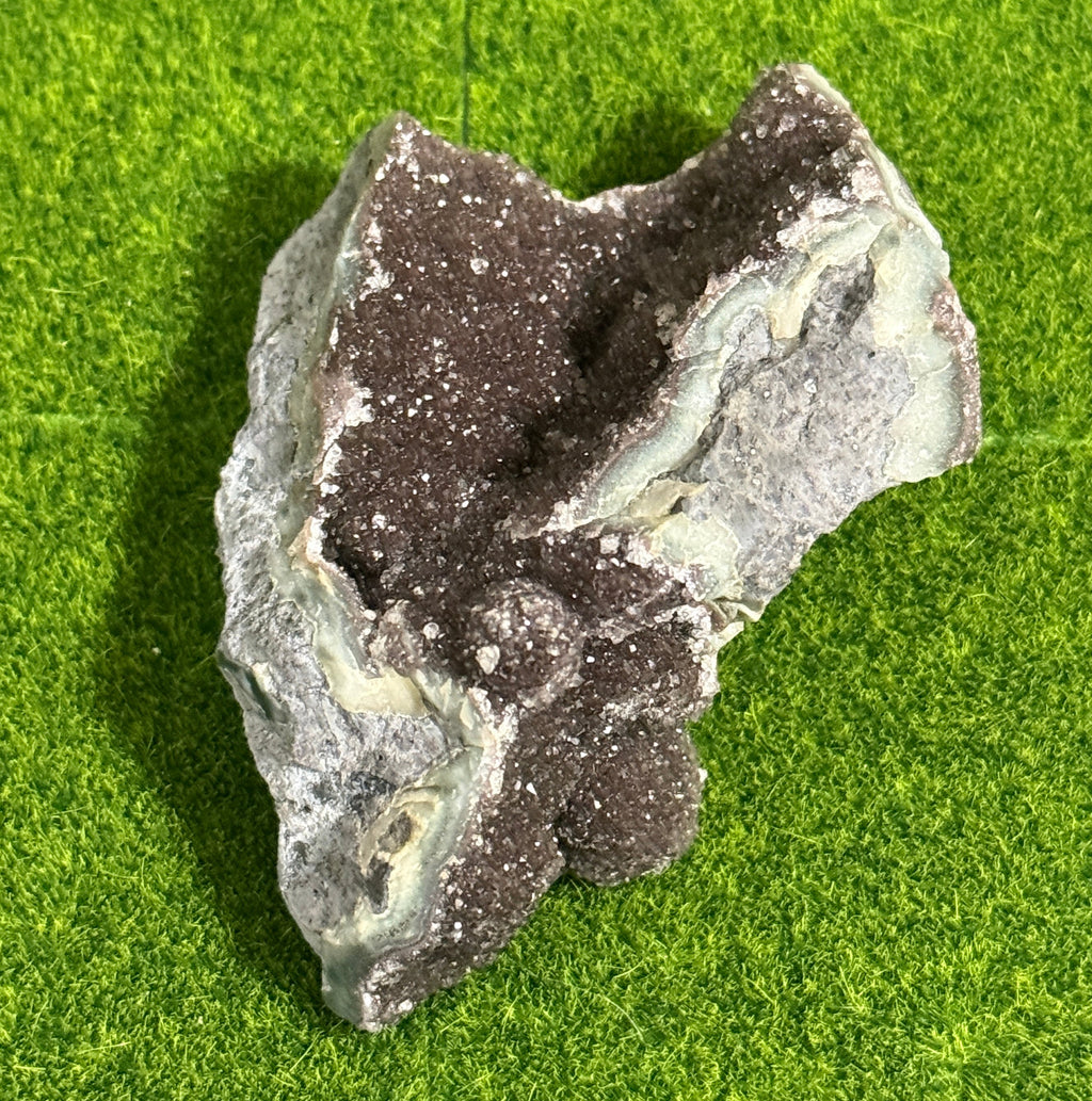 A natural, irregularly shaped rock formation with a dark brown, crystal-like interior and a gray and light green exterior. The rock sits on a green surface, showcasing its unique texture and color variations.