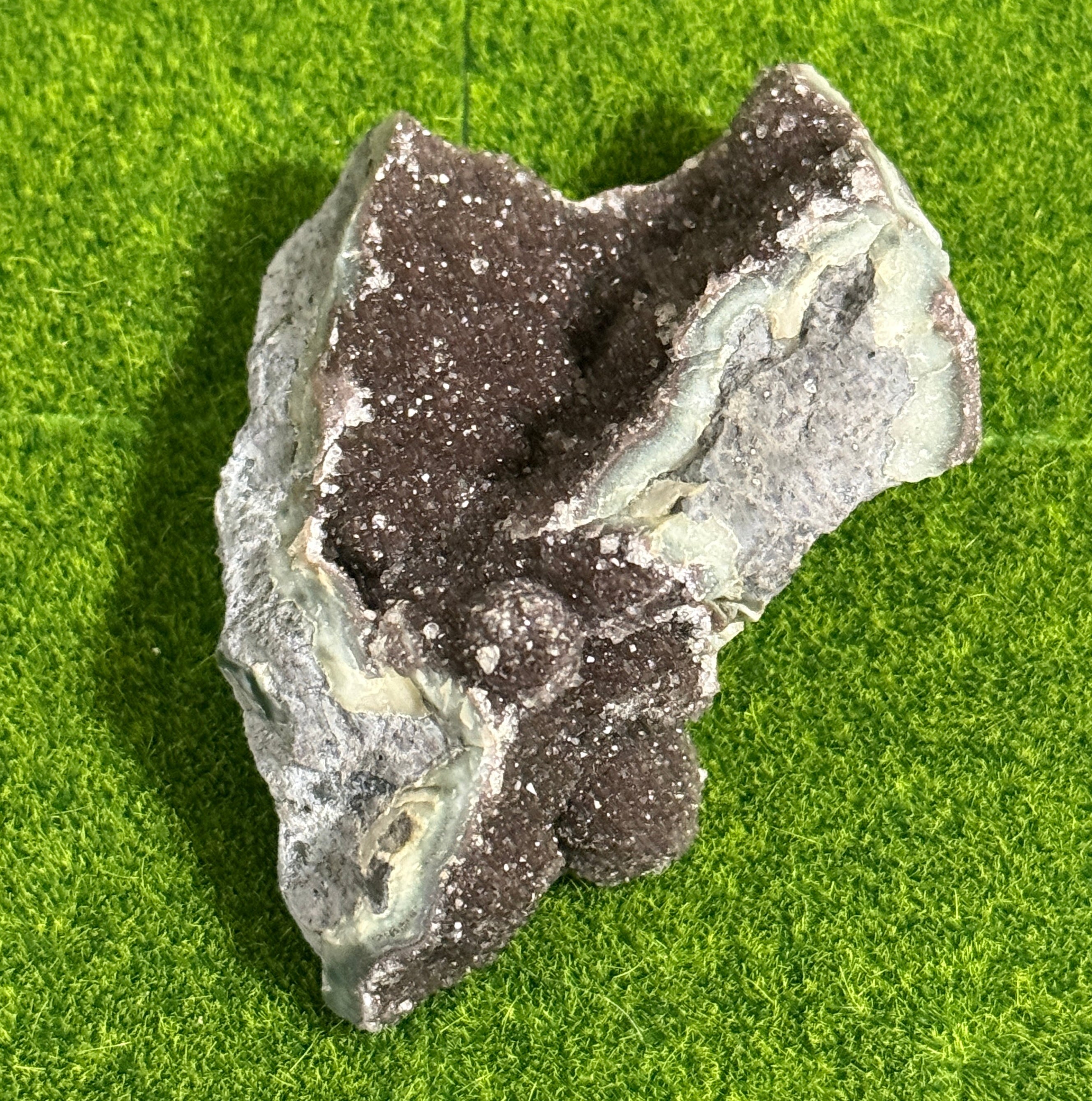 A natural, irregularly shaped rock formation with a dark brown, crystal-like interior and a gray and light green exterior. The rock sits on a green surface, showcasing its unique texture and color variations.