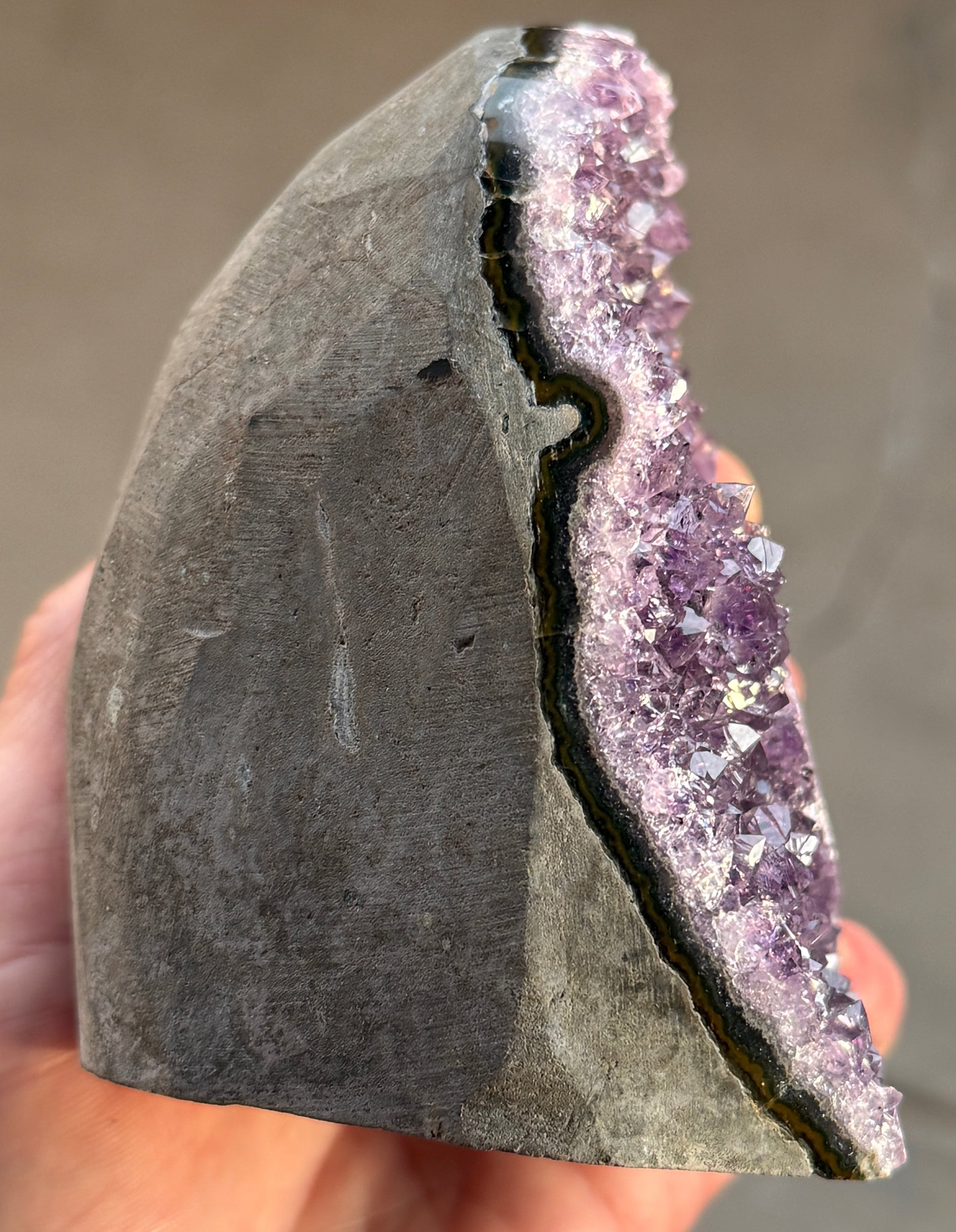 Side view of a natural amethyst geode with a deep purple crystal interior. The geode has a rounded shape and a rough, textured exterior. A small, circular formation with green and yellow hues is visible within the crystal cluster.