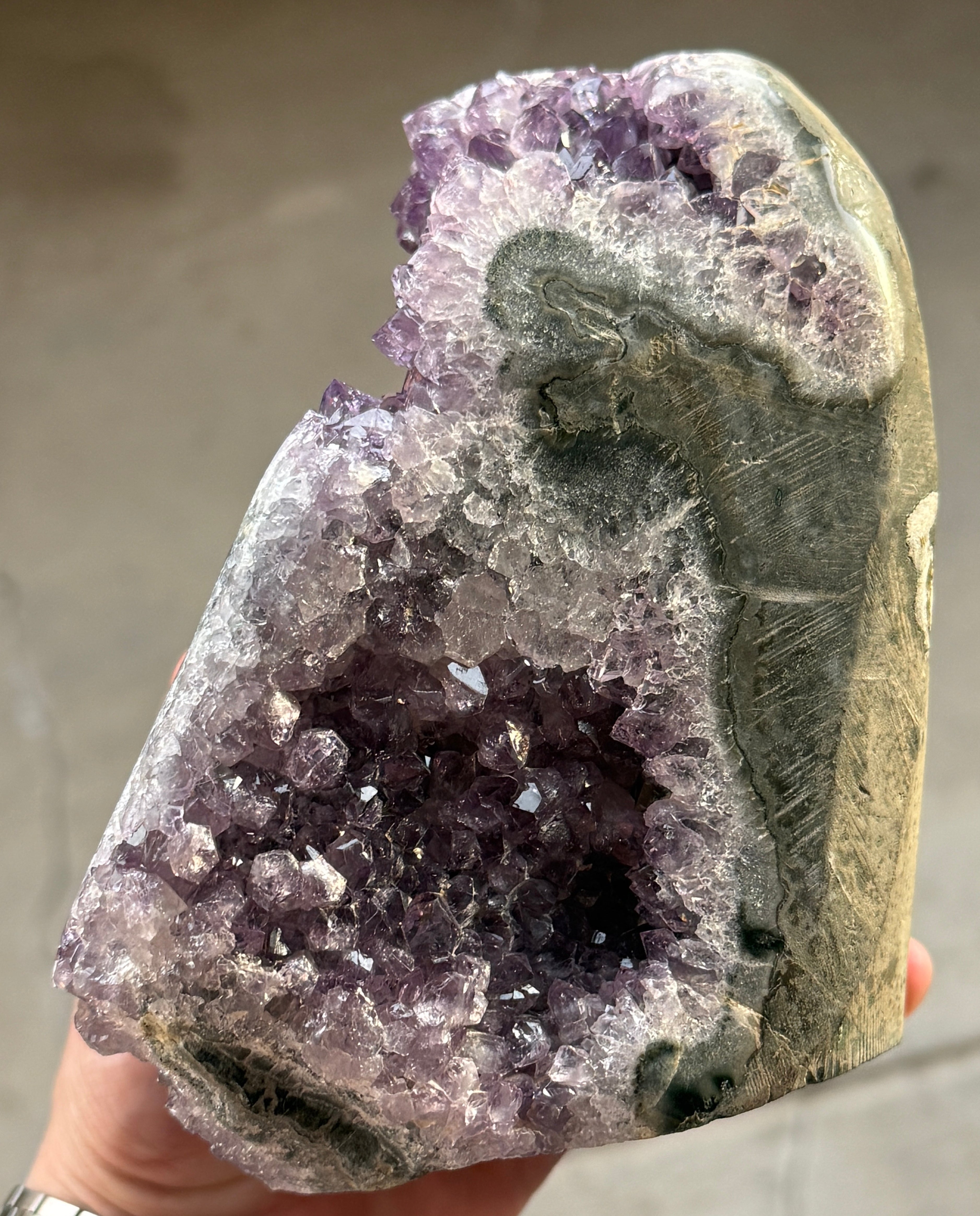 A large amethyst geode with a deep purple crystalline interior. The geode has a rough, natural exterior with a gray and white border. The crystals are densely packed and reflect light, creating a sparkling effect.