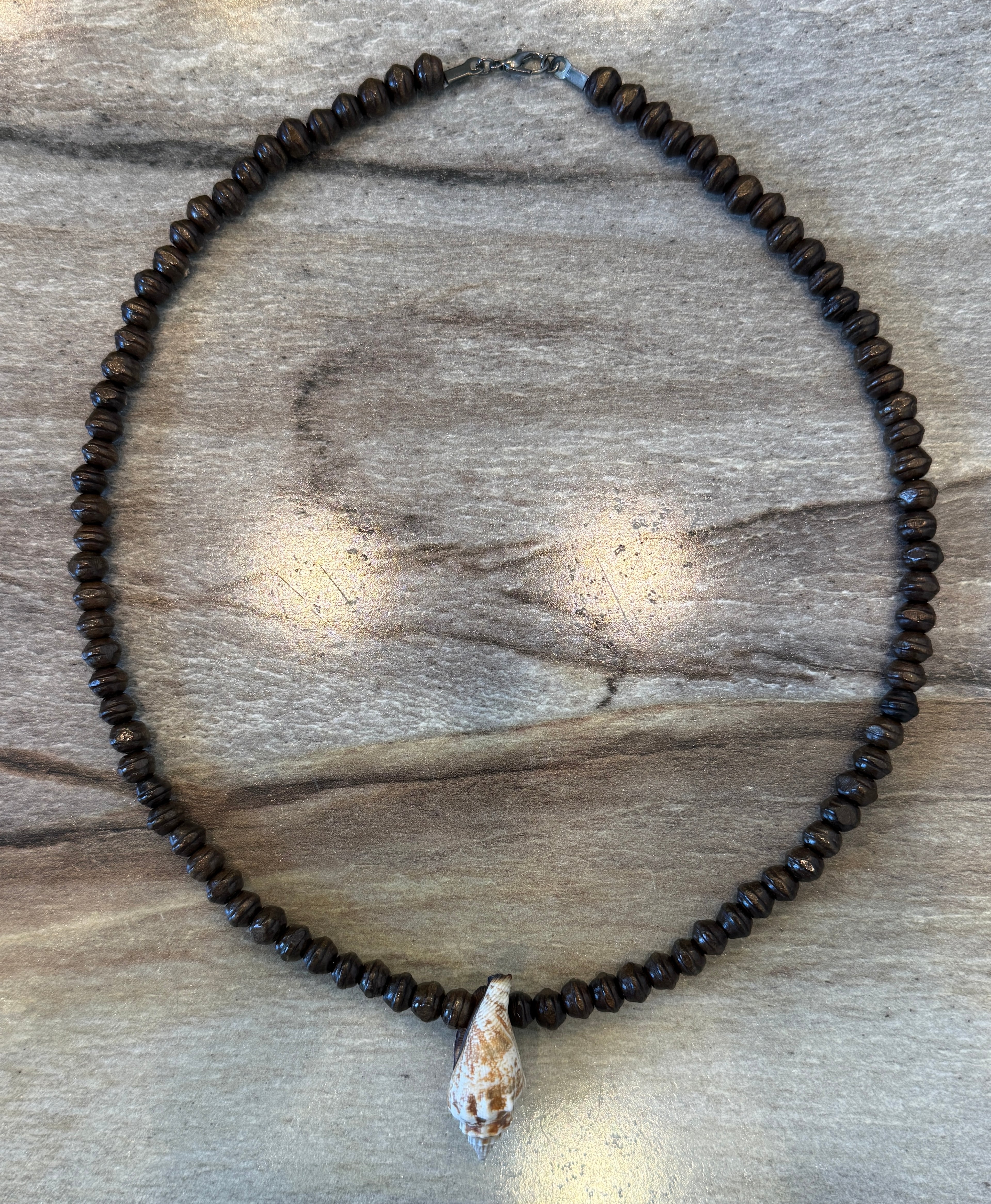 A beaded necklace made of small dark brown beads with a white seashell pendant, displayed on a gray wood background.