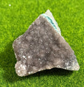 A rough-edged, triangular-shaped crystal cluster. The crystal has a dark gray, speckled surface with a lighter, crystalline edge and a turquoise-colored interior. The crystal sits on a bright green surface.