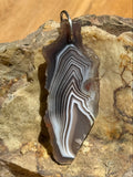 A polished agate stone pendant with concentric banding in black, gray, white, and brown, featuring a small metal loop at one end, displayed on a textured rock surface.