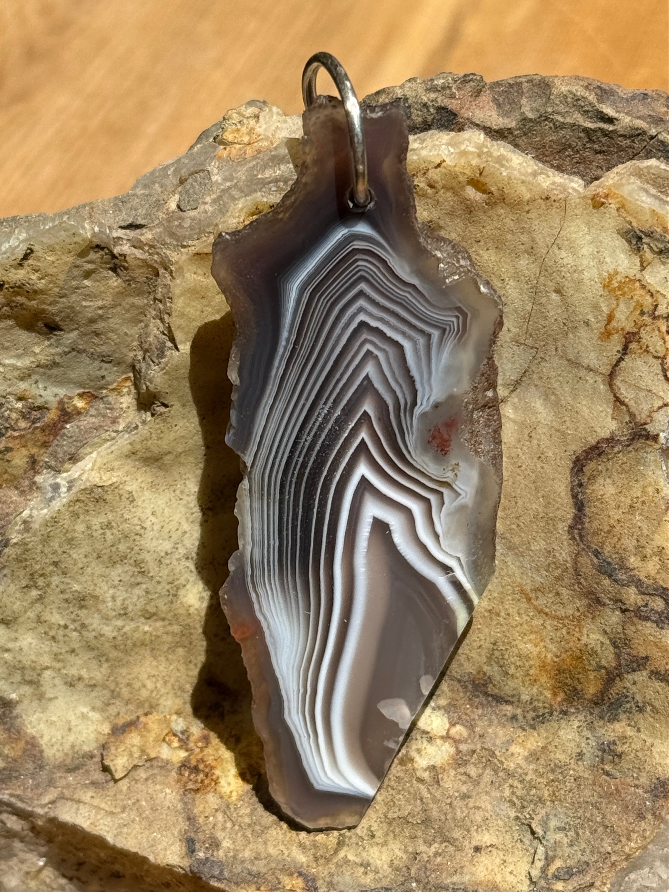 A polished agate stone pendant with concentric banding in black, gray, white, and brown, featuring a small metal loop at one end, displayed on a textured rock surface.