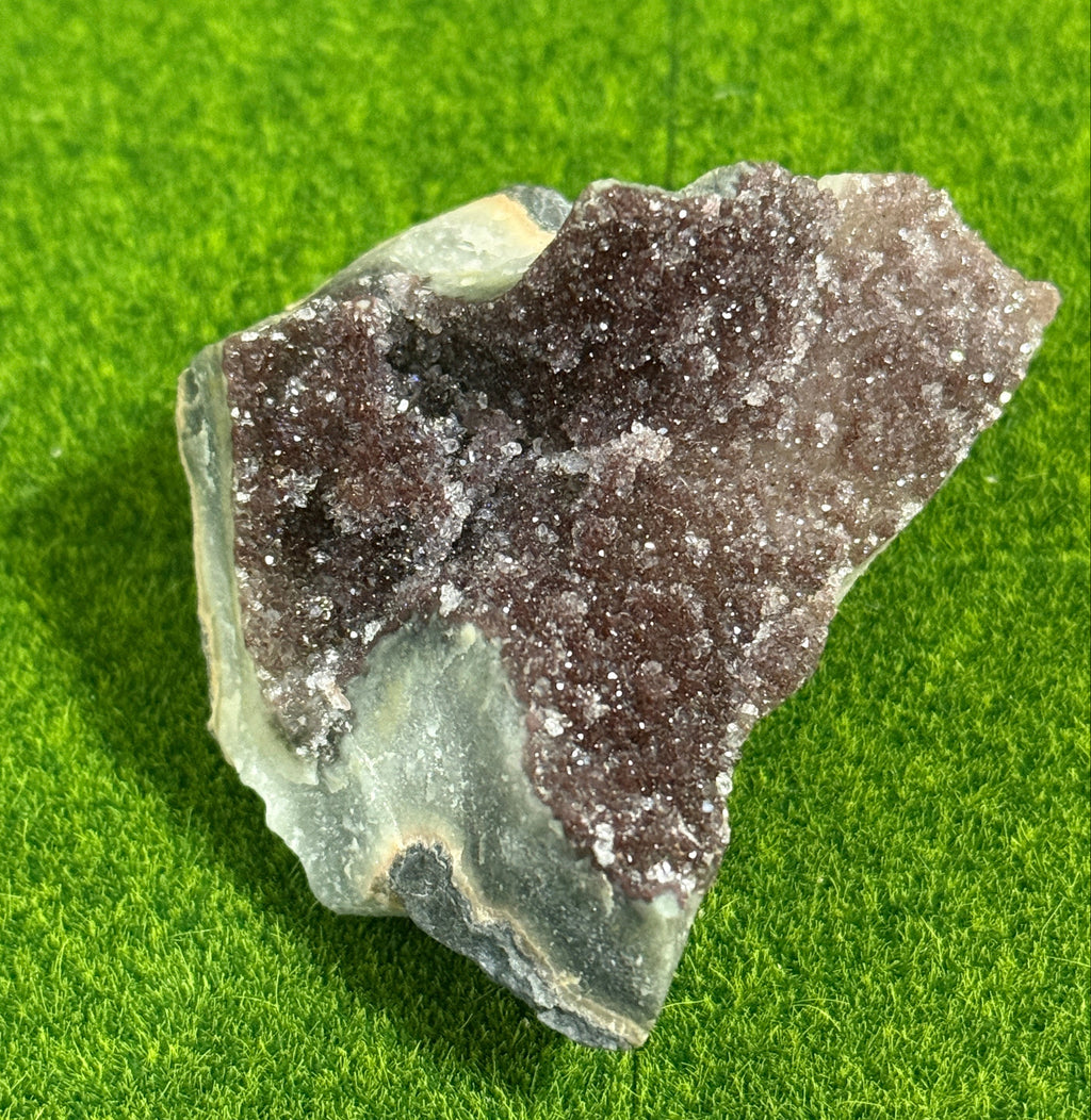 A raw amethyst geode with a dark purple crystalline interior and a light green and gray exterior. The geode is set against a green background.