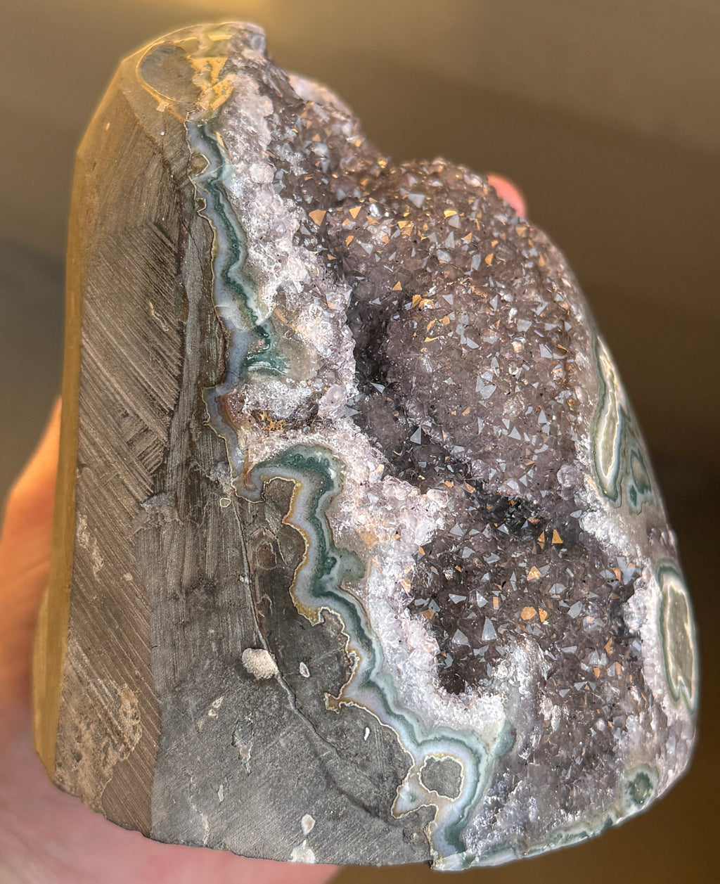 A large, oval-shaped geode with a dark purple interior filled with sparkling crystals. The outer shell is a mottled gray-green with areas of teal and gold. The geode sits on a white surface.